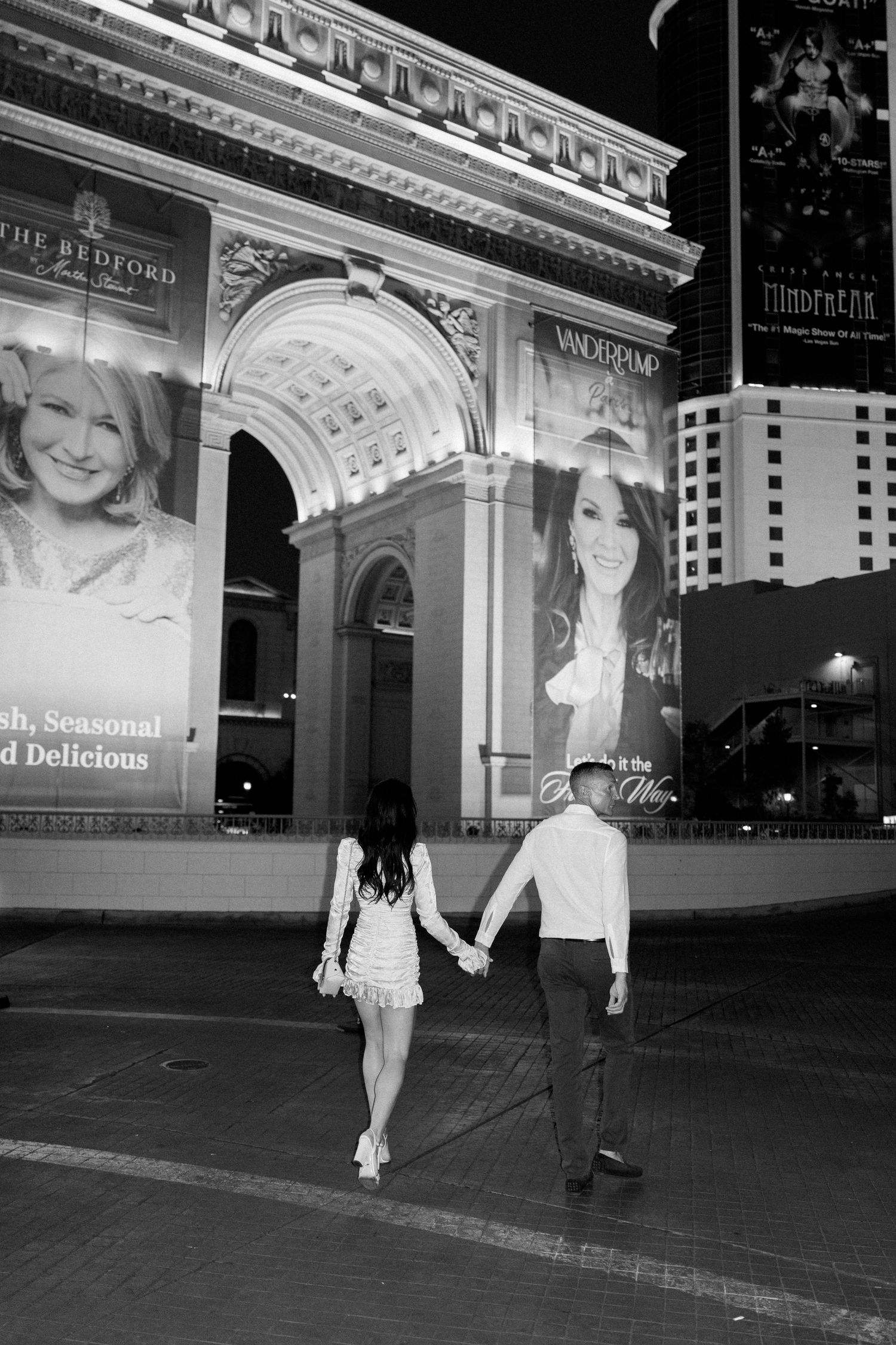 A black and white photo of a couple walking past an illuminated archway with advertising billboards.