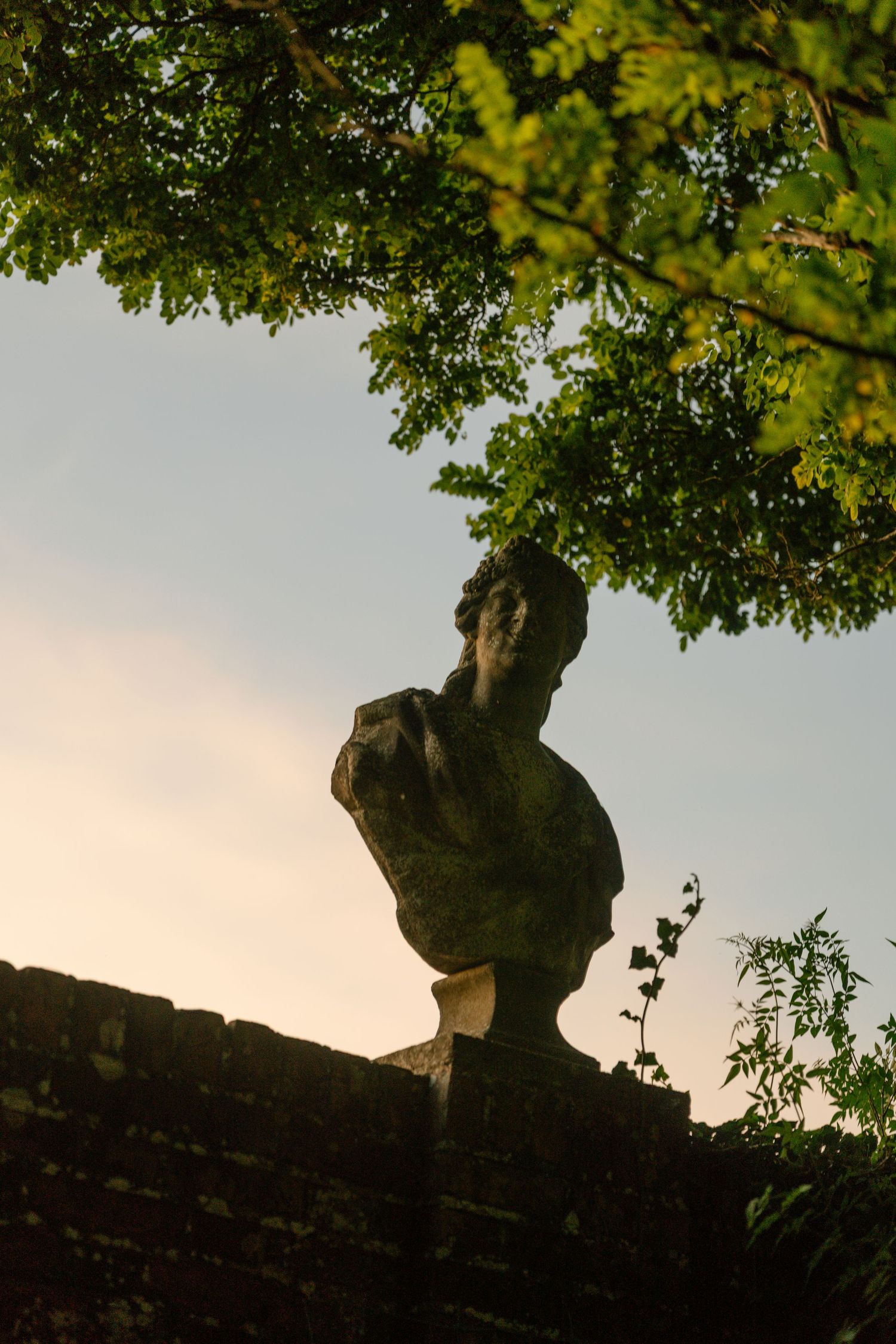 Stone statue silhouetted against sunset sky with tree branches overhead.