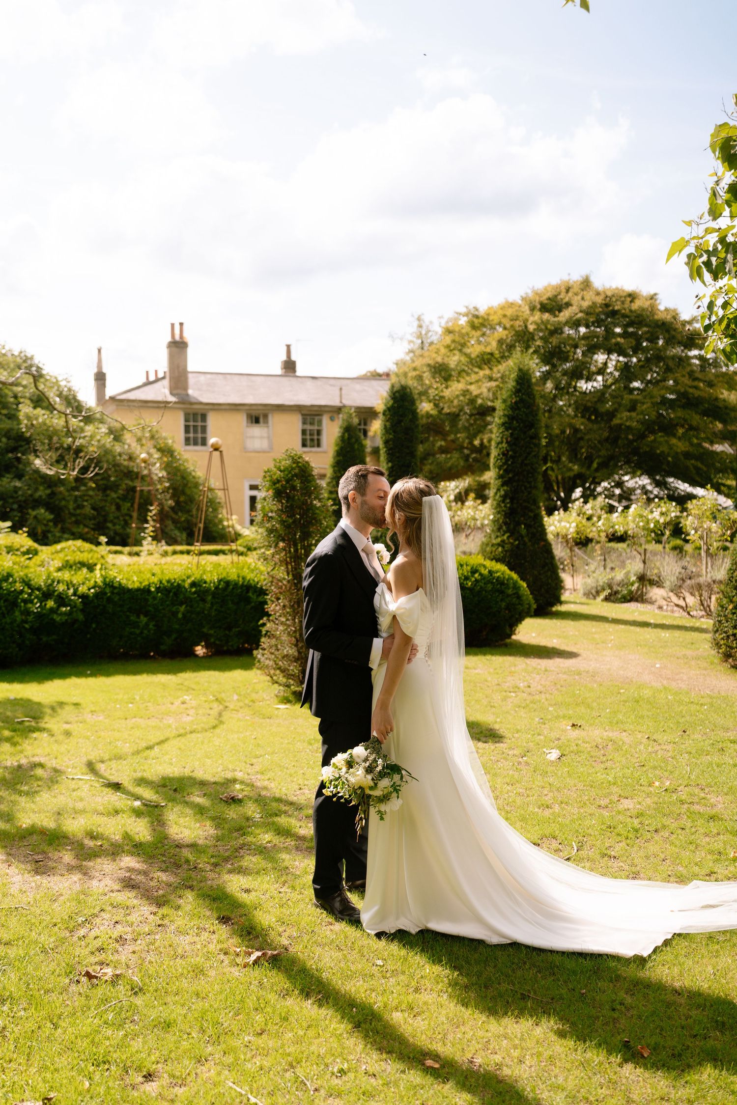 Wedding couple poses on manicured lawn with elegant yellow manor house in background.
