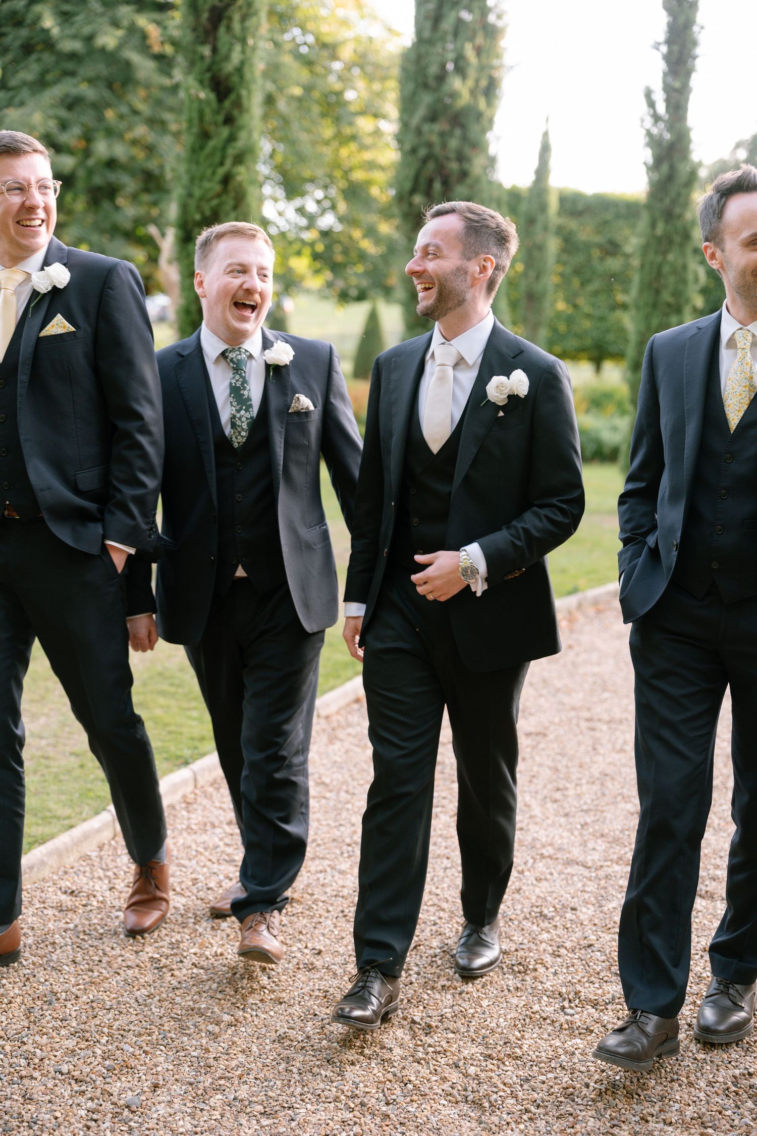 Candid moment of groomsmen laughing while walking on a garden path in formal attire.