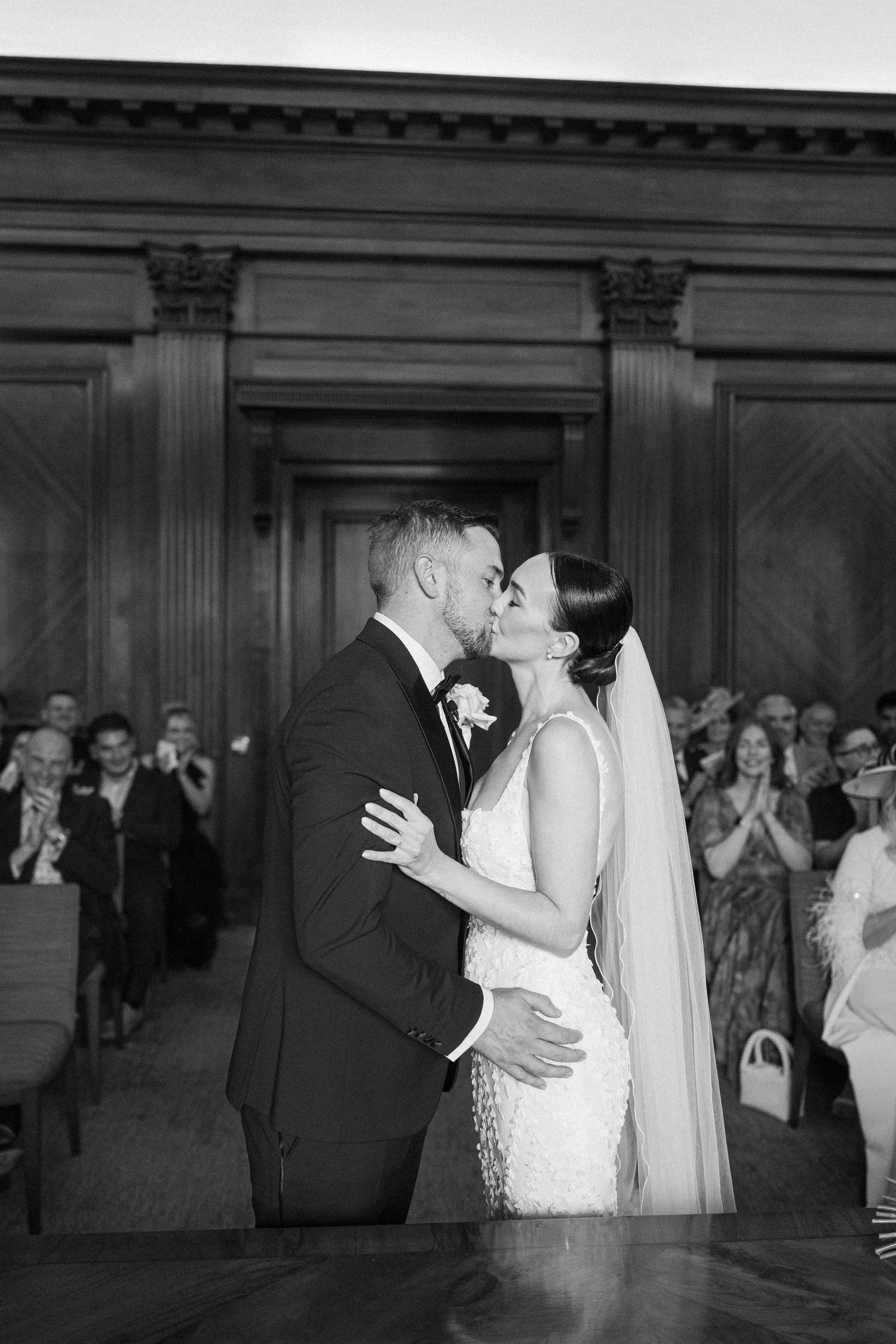 A romantic wedding kiss captured in black and white with guests applauding in the background.