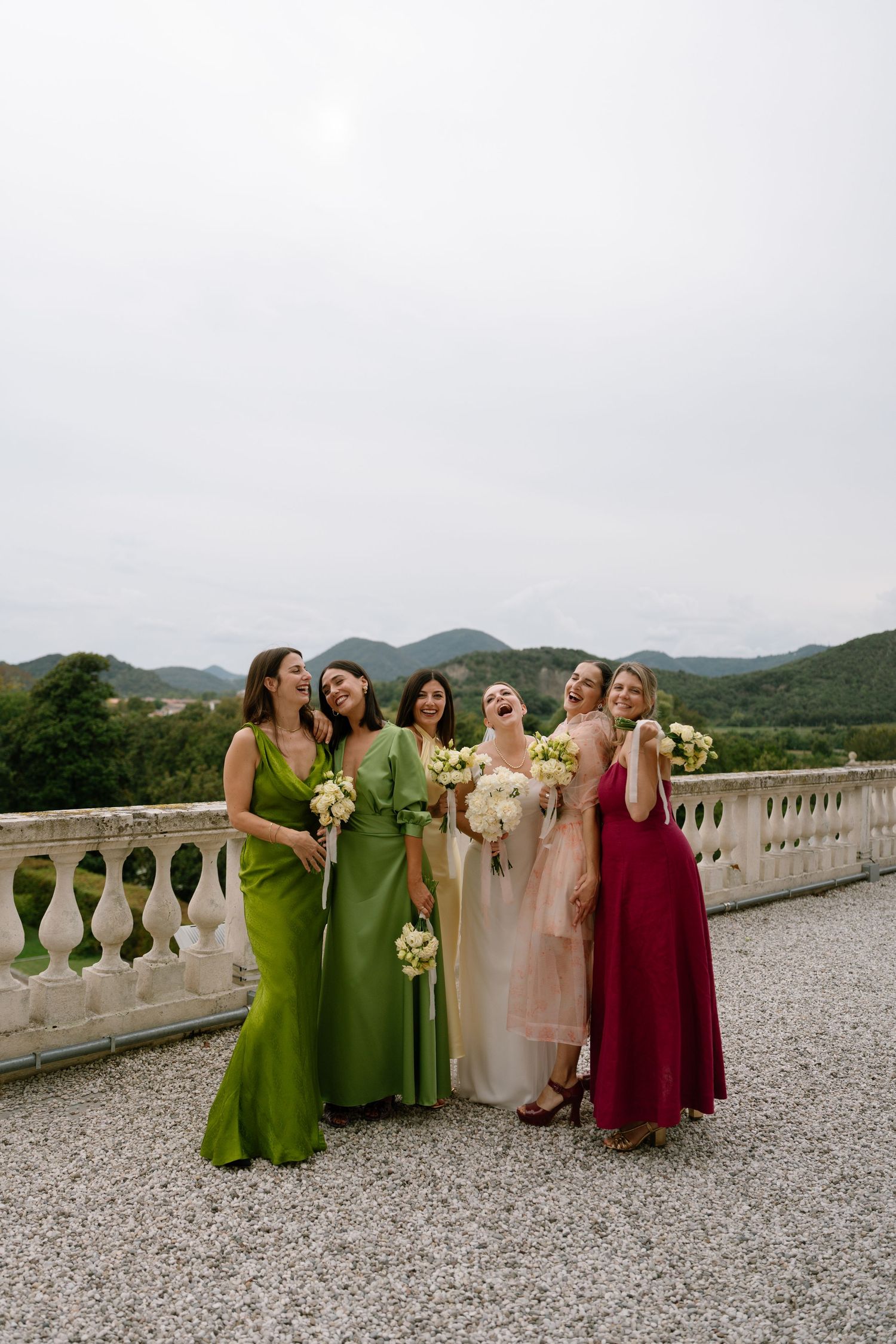 Bridal party in jewel-toned dresses stands together on a stone terrace with mountain views.