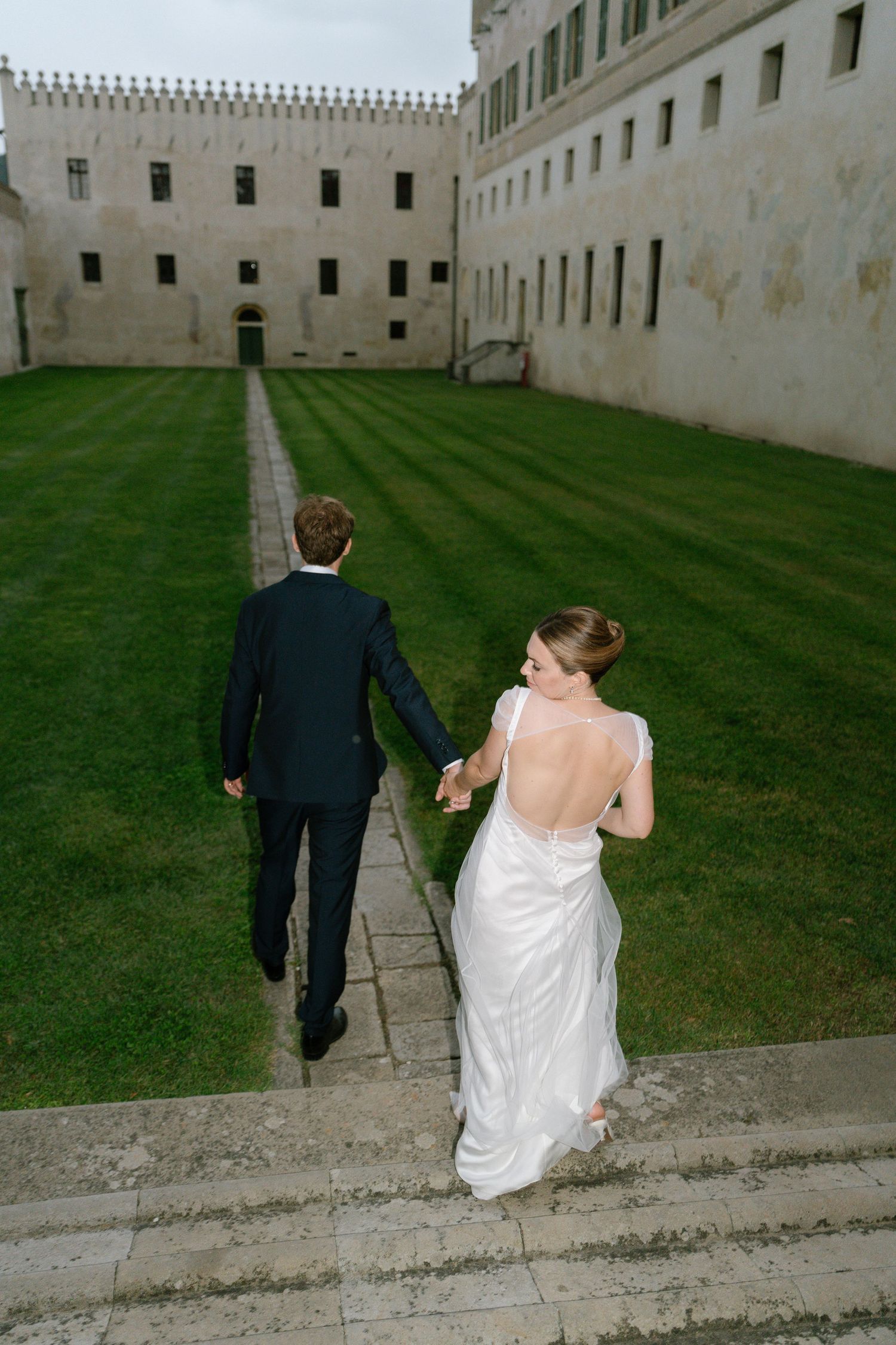Couple walks along a grassy path beside a historic stone building with white walls.