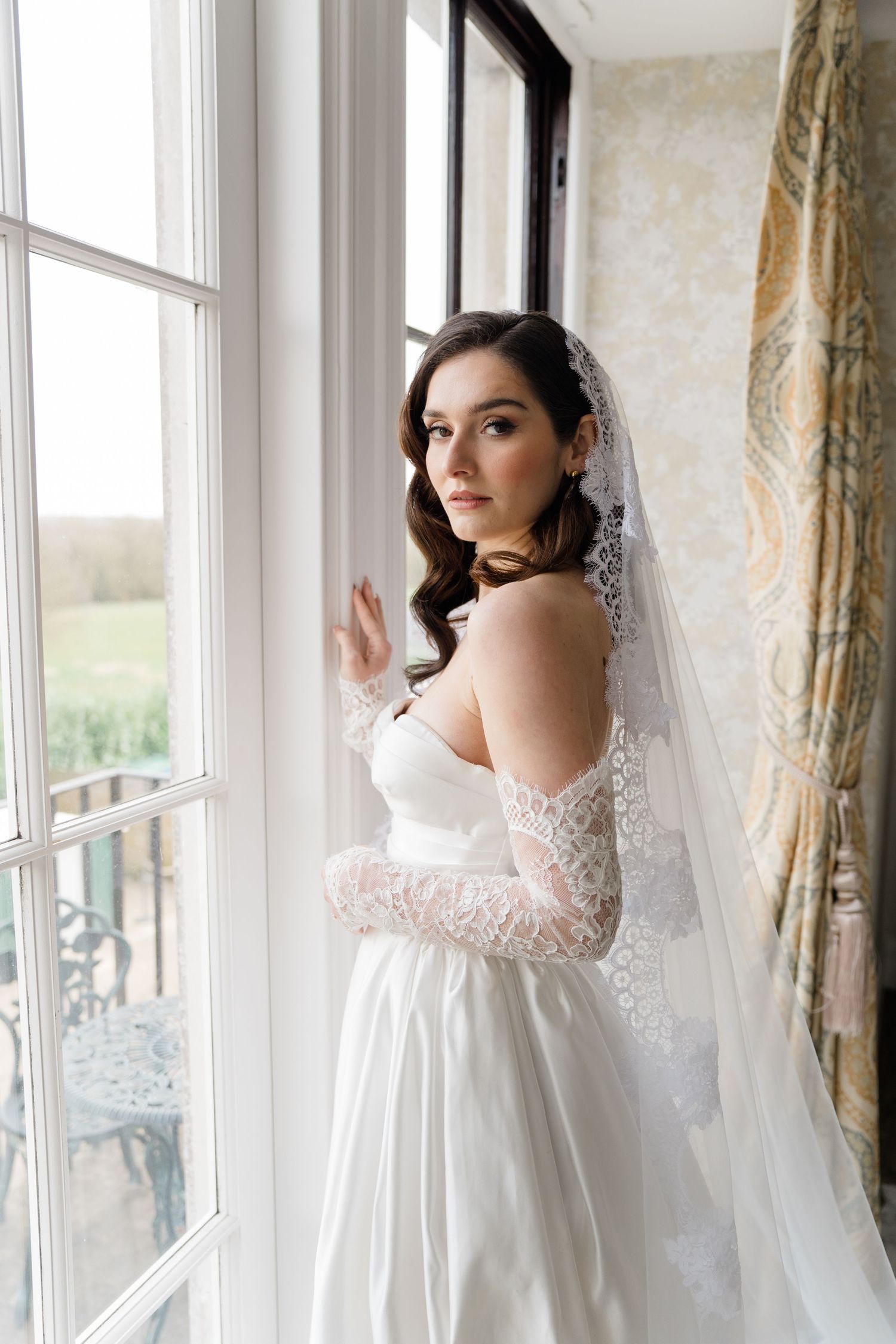 Bride in white lace gown poses by window in romantic bridal portraits.