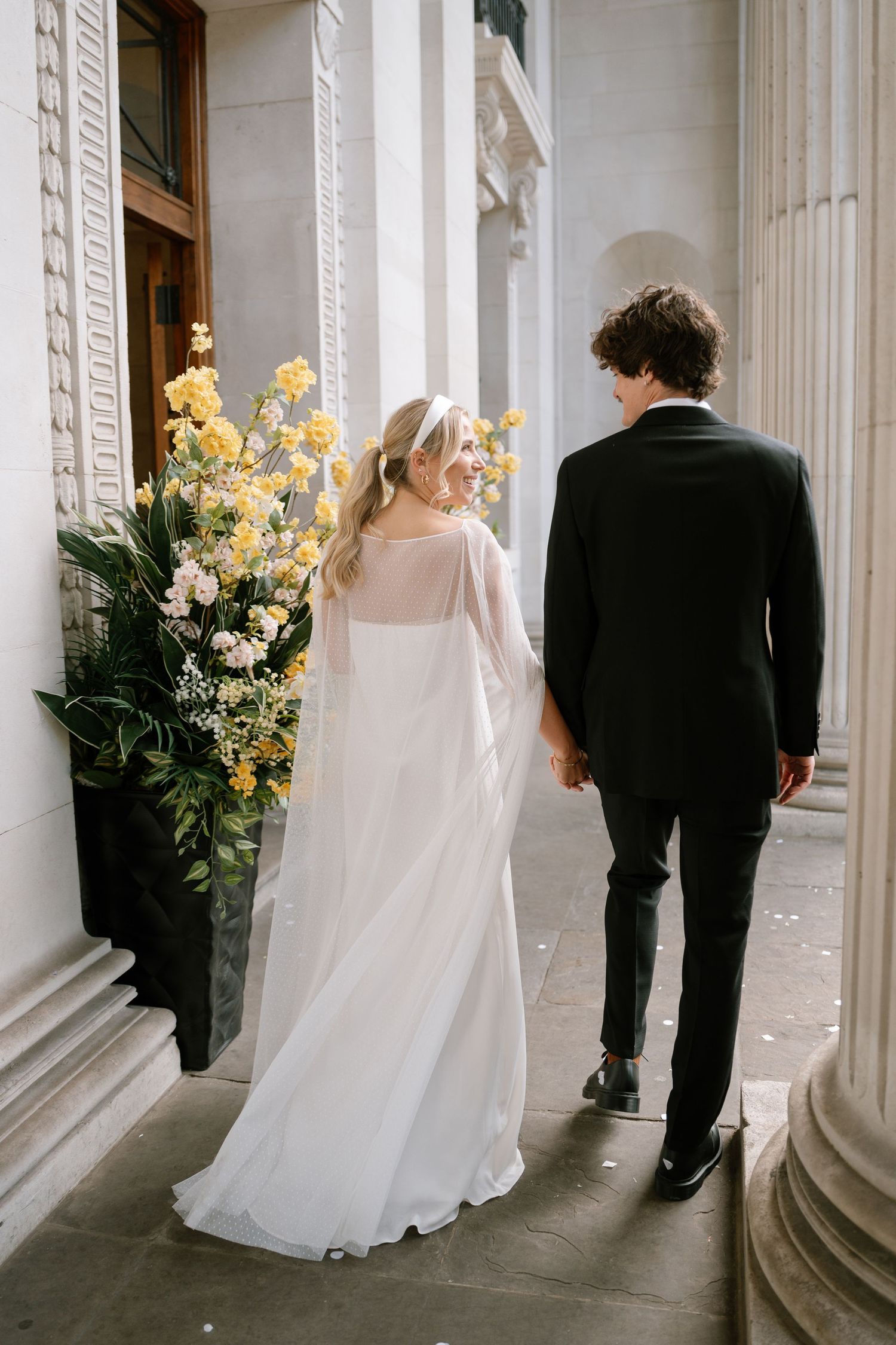 A wedding couple walking hand in hand past classical architecture and yellow floral arrangements.