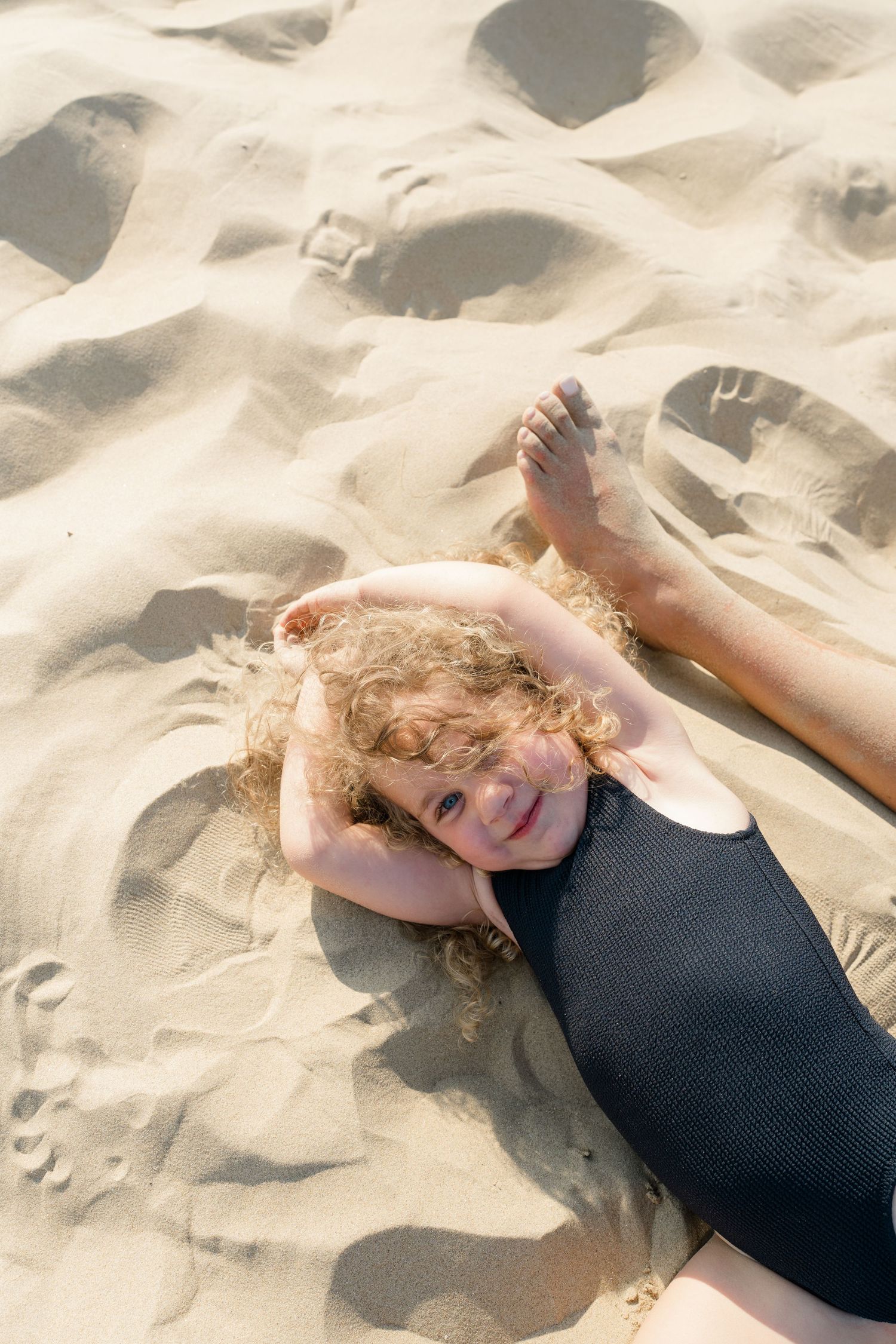 Person with curly hair resting on sandy beach in black swimsuit.