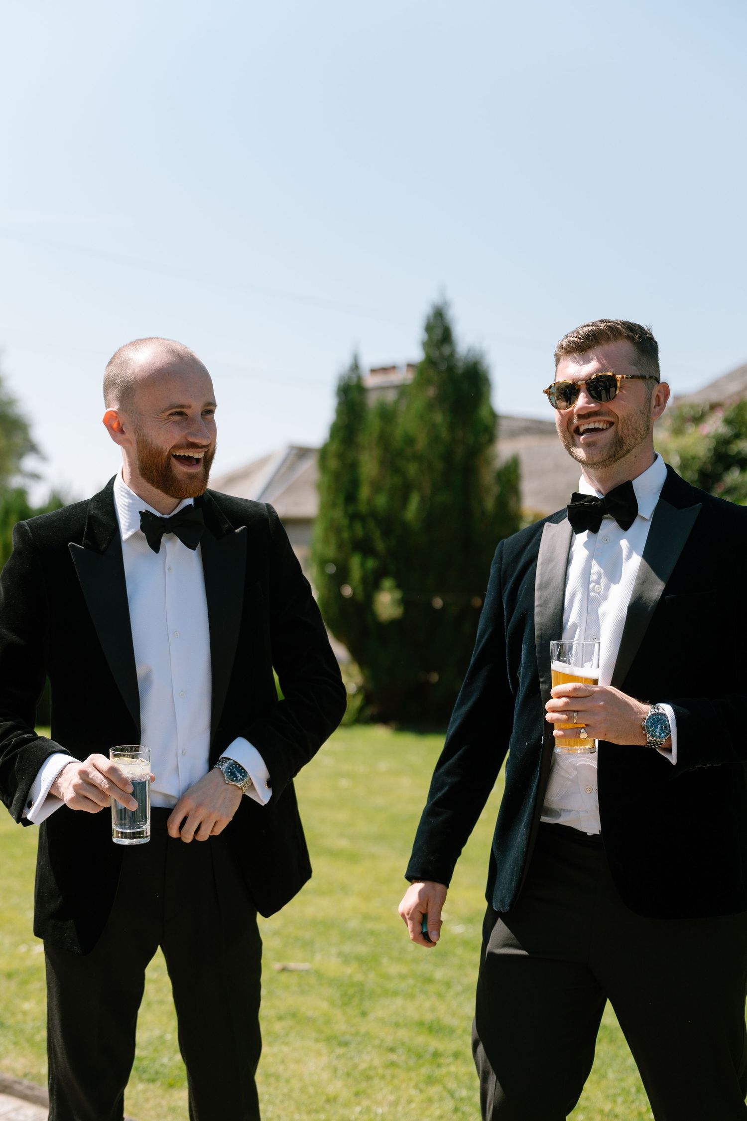 Men in tuxedos sharing drinks and laughs on a sunny lawn.