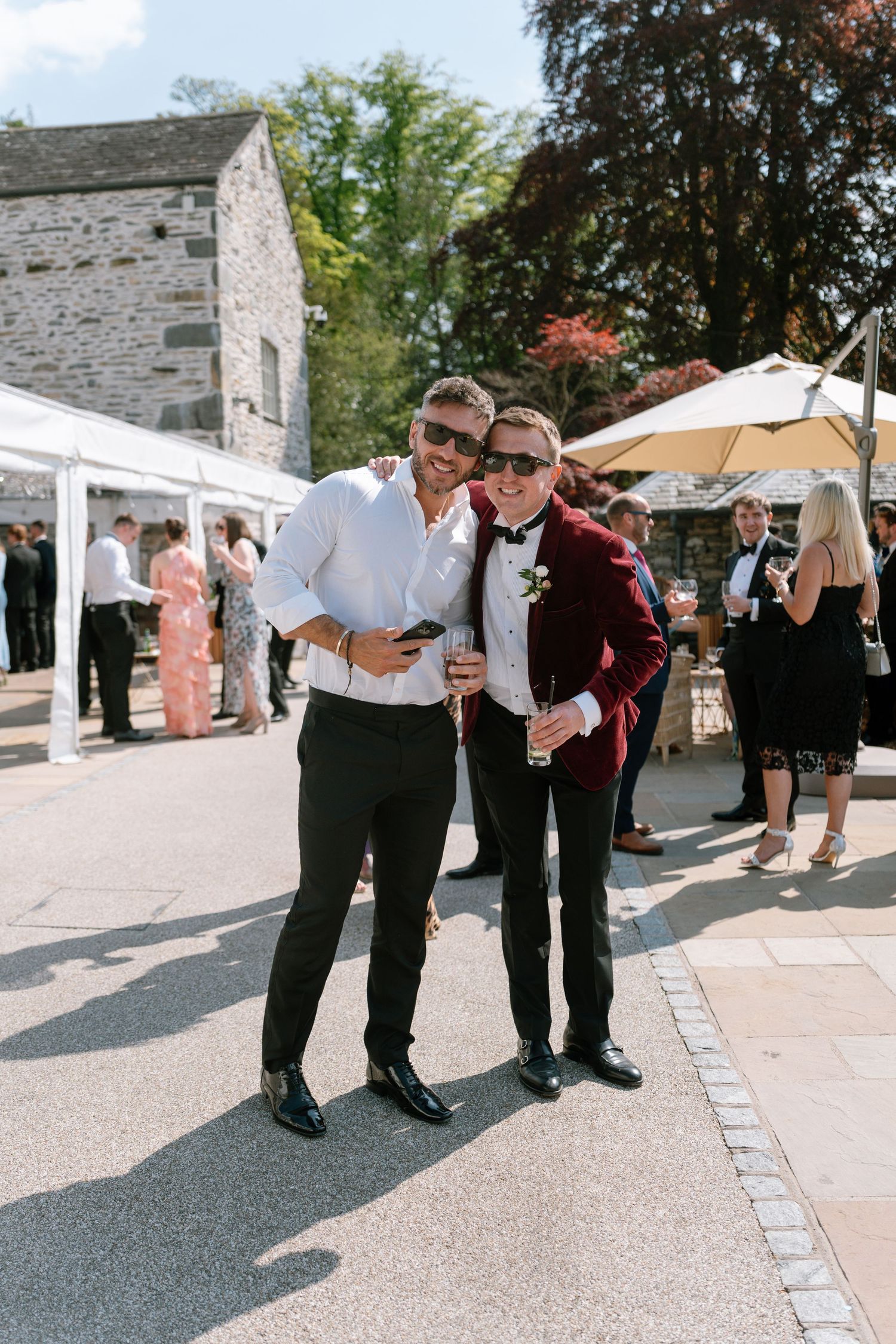 Wedding guests in formal attire gather outside a stone building on a sunny day.