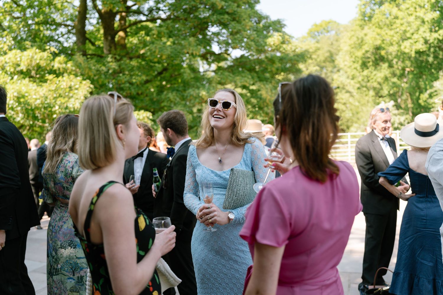 Wedding guests gather outdoors on a sunny day surrounded by lush green trees.