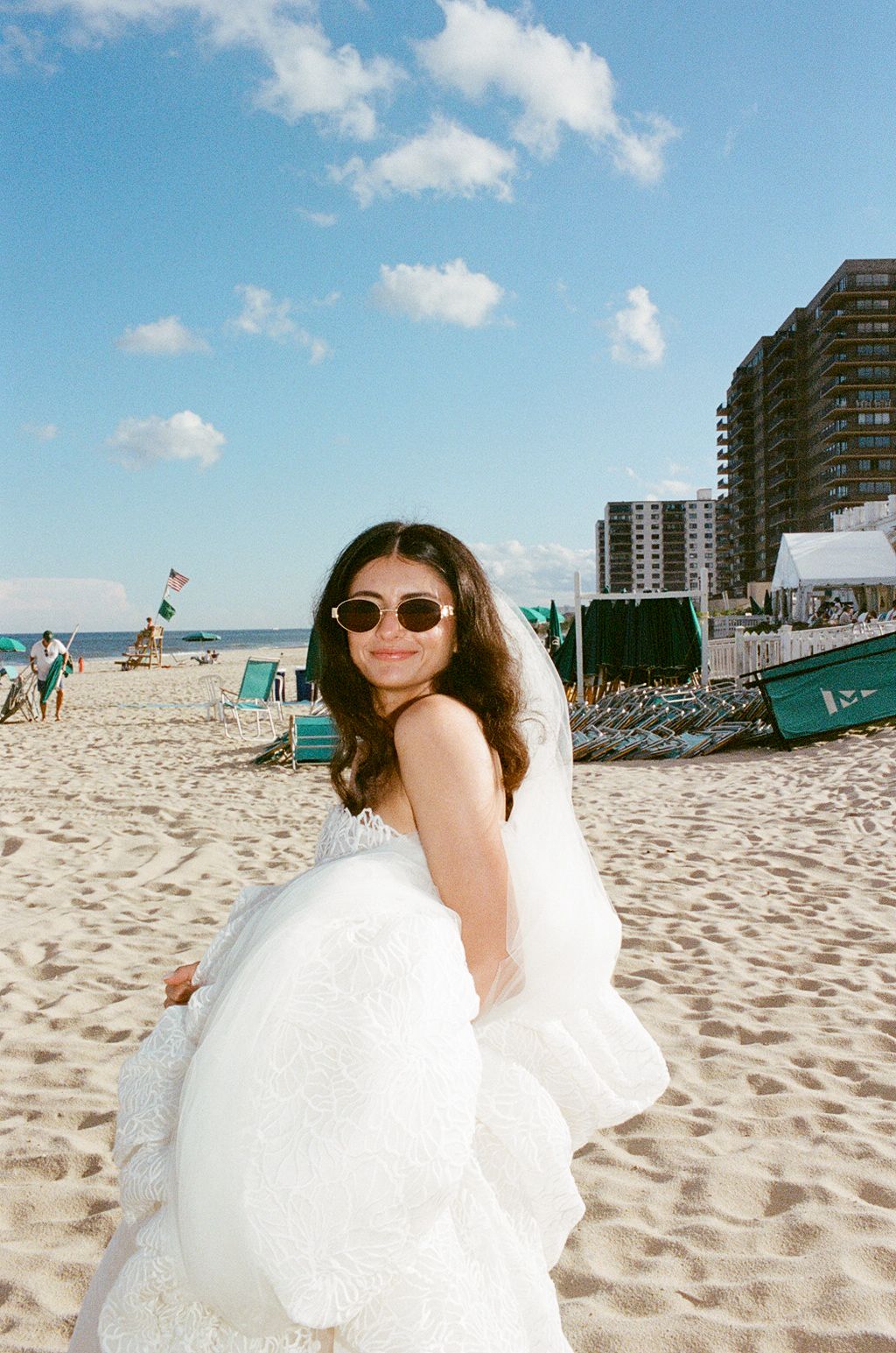 Bride in white wedding gown and sunglasses smiling on a sunny beach with buildings and blue sky.