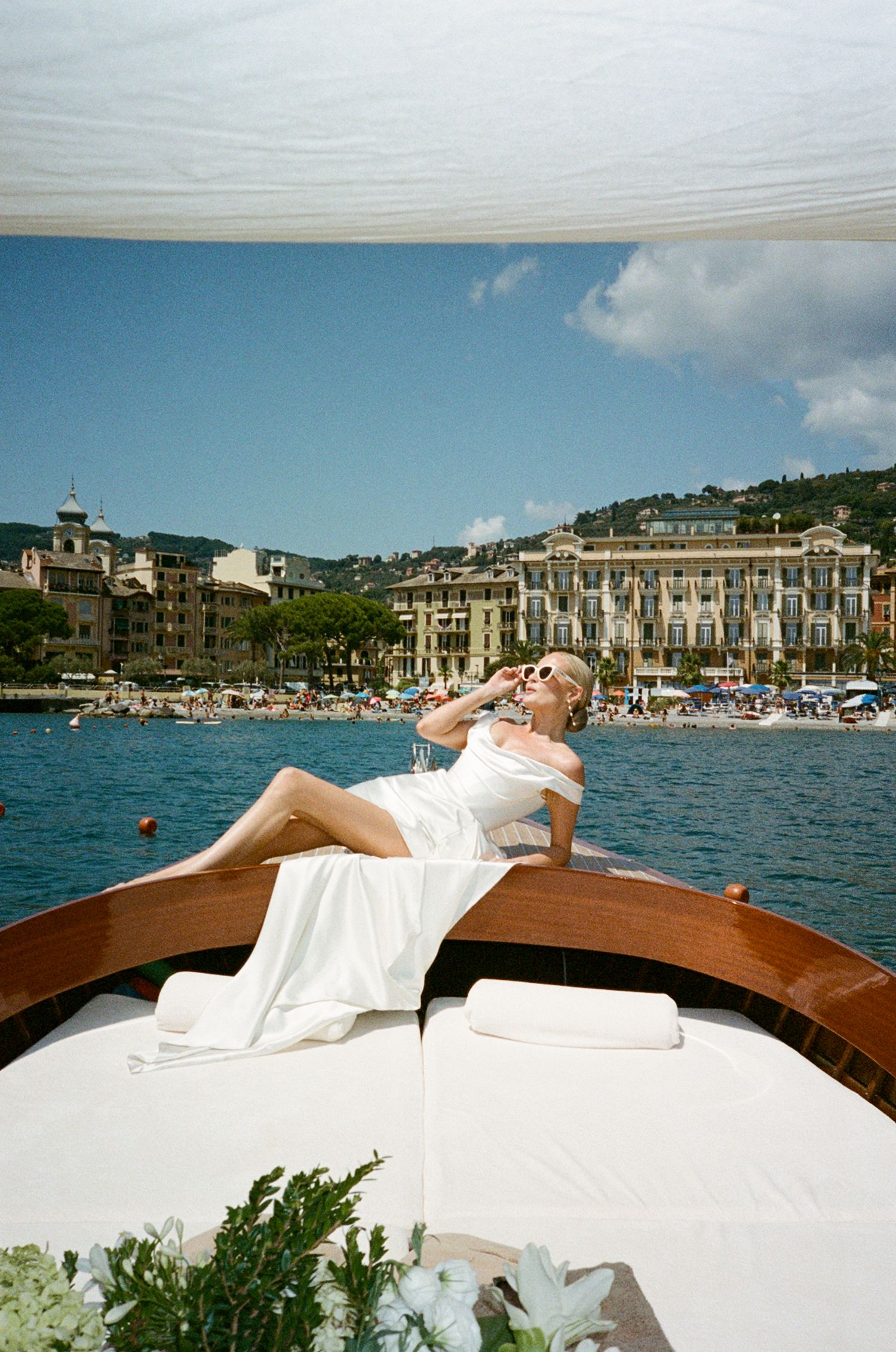 Woman in white dress lounging on luxury boat bow with scenic Italian coastal town and blue sky backdrop.