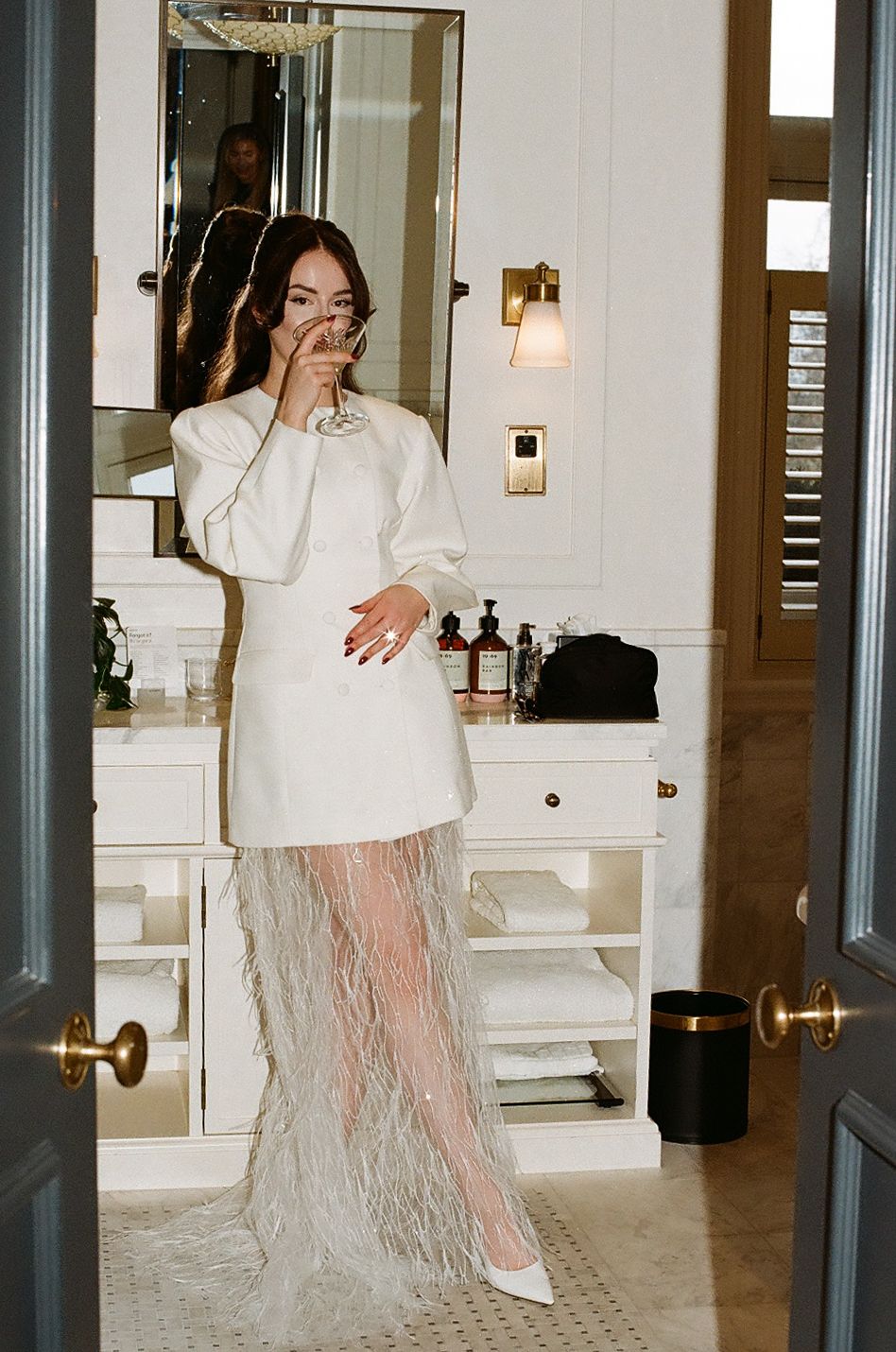 A person in a white oversized top and sheer feathered skirt poses in an elegant bathroom interior with marble details.