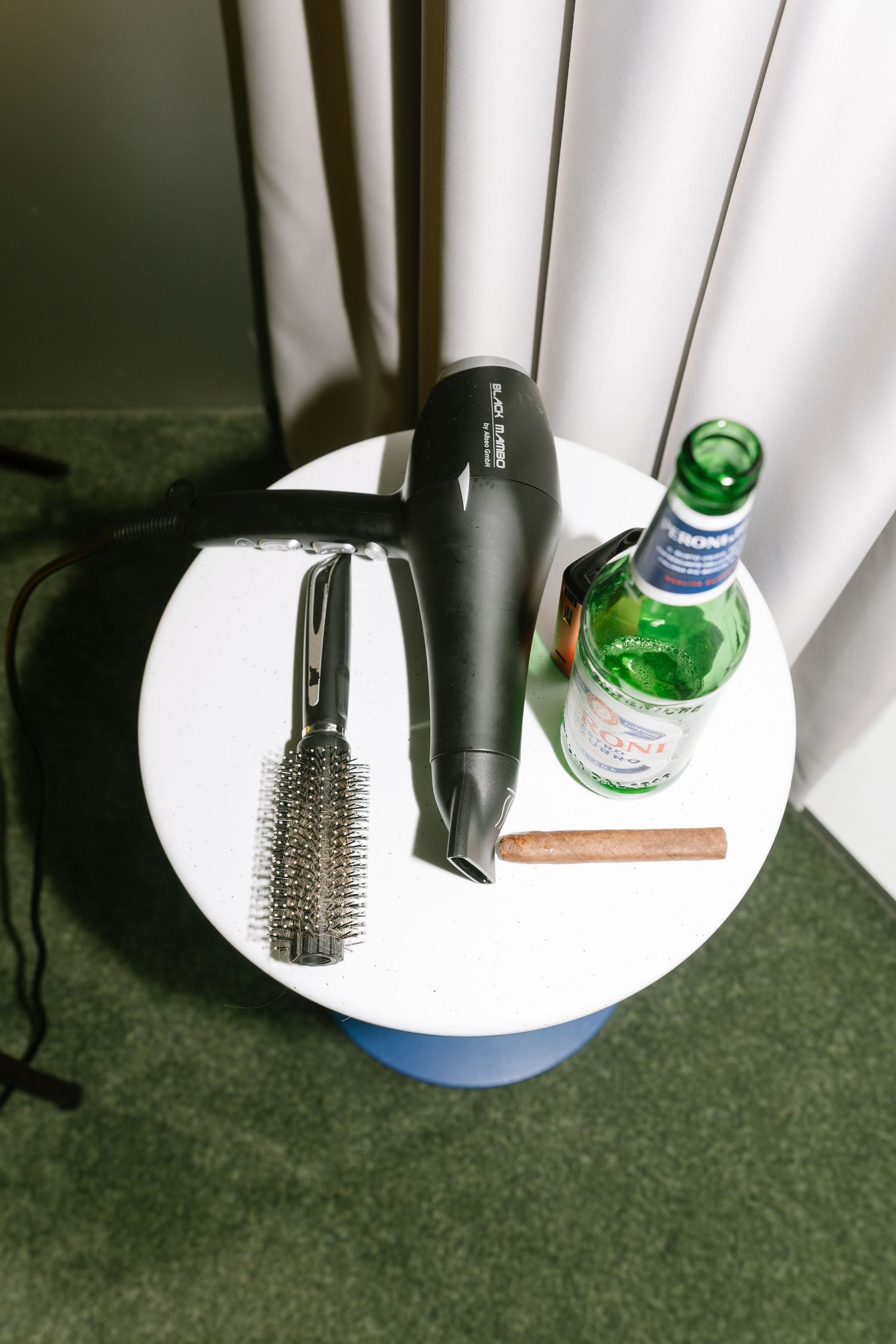Hair styling tools and beverage arranged on white side table with curtain backdrop.