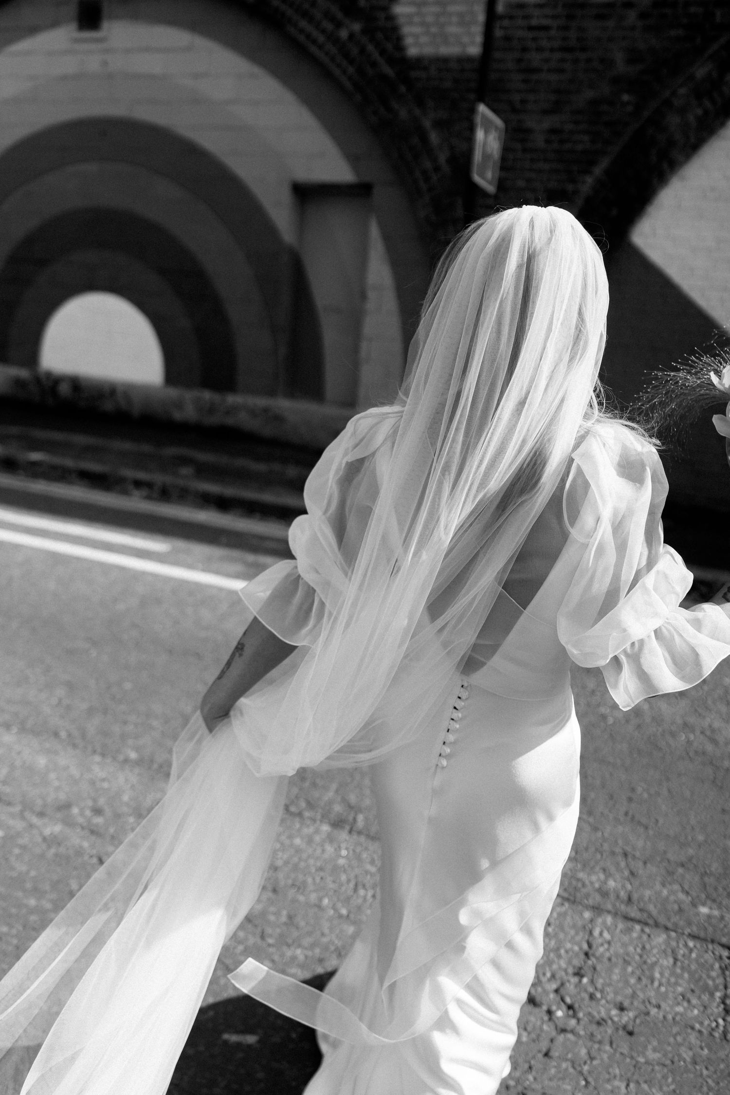 A bride in a white wedding dress with a flowing veil stands under an arched passageway in black and white.