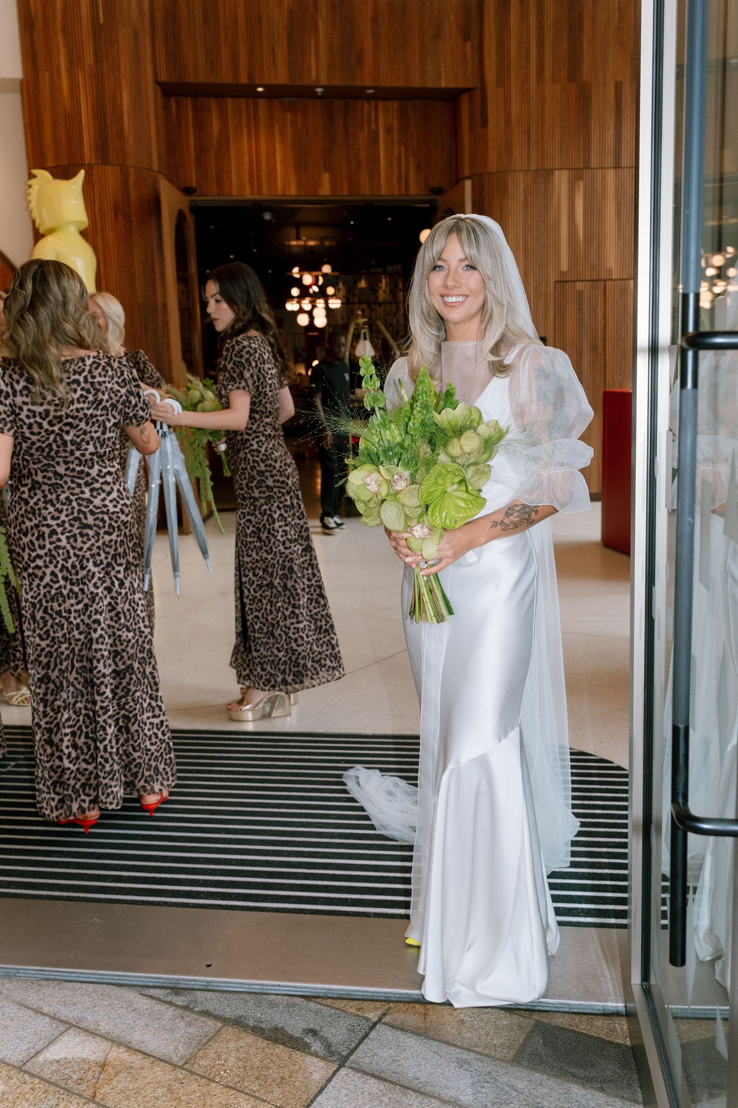 Wedding party in white gowns and leopard print dresses walking through modern venue entrance.