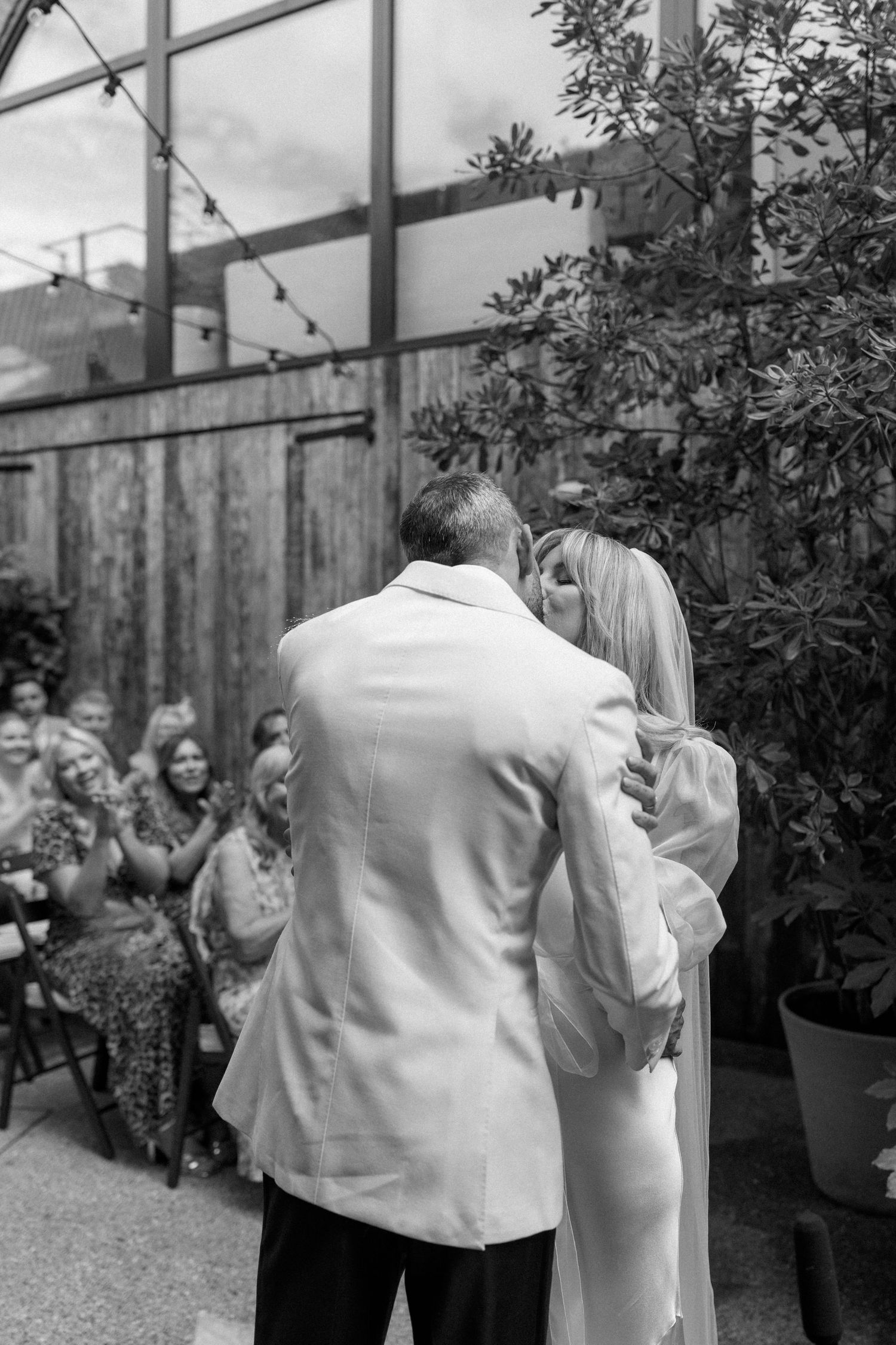 Wedding ceremony guests gathering in an outdoor greenhouse space with potted plants.