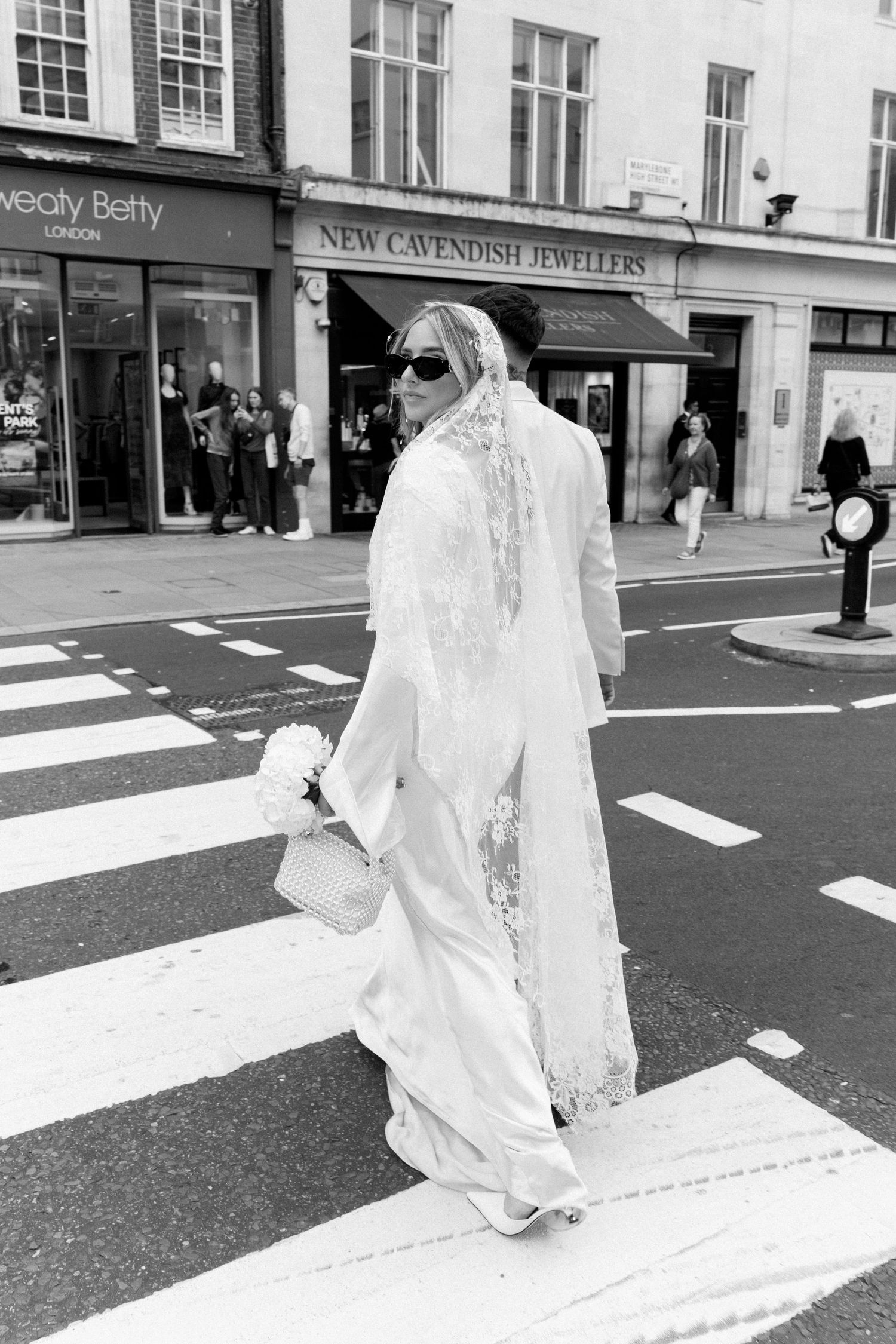 A bride in a flowing white dress and veil crosses a London street near shops.