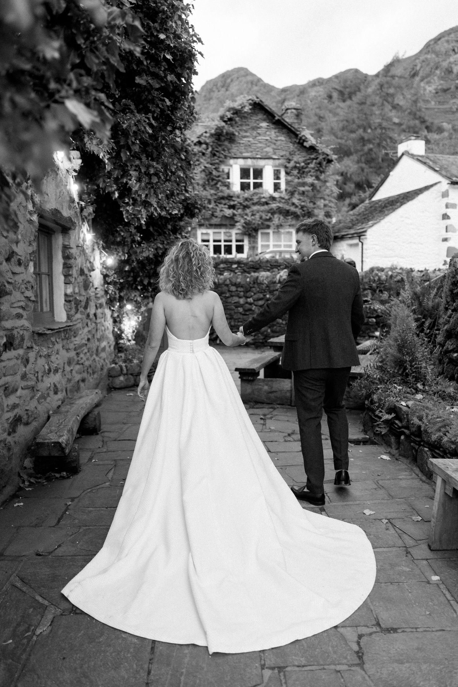 Wedding couple walks along a stone path with mountains and houses in the background.