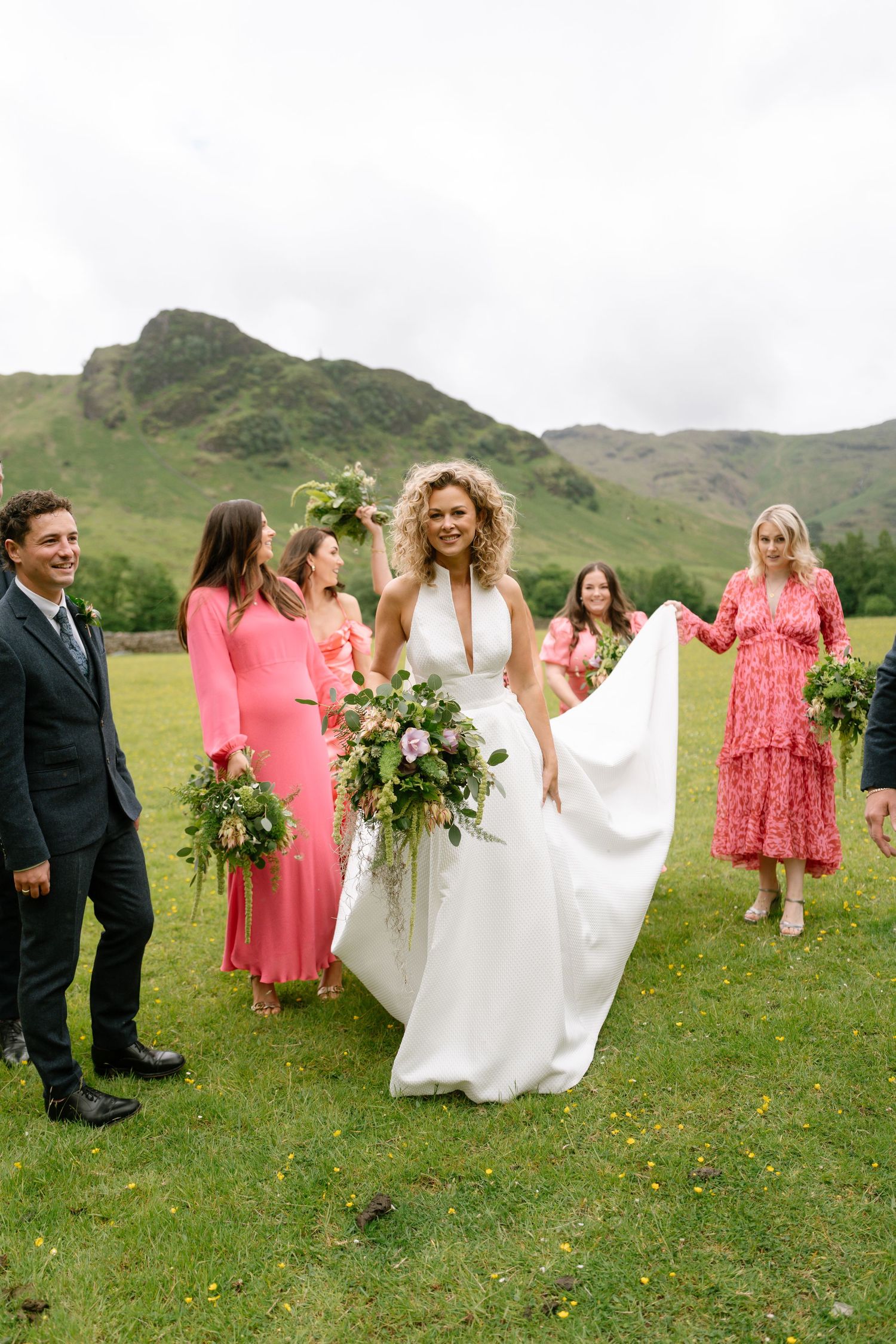 Wedding party poses on a green hillside with bridesmaids in pink dresses and a bride in white holding greenery bouquets.
