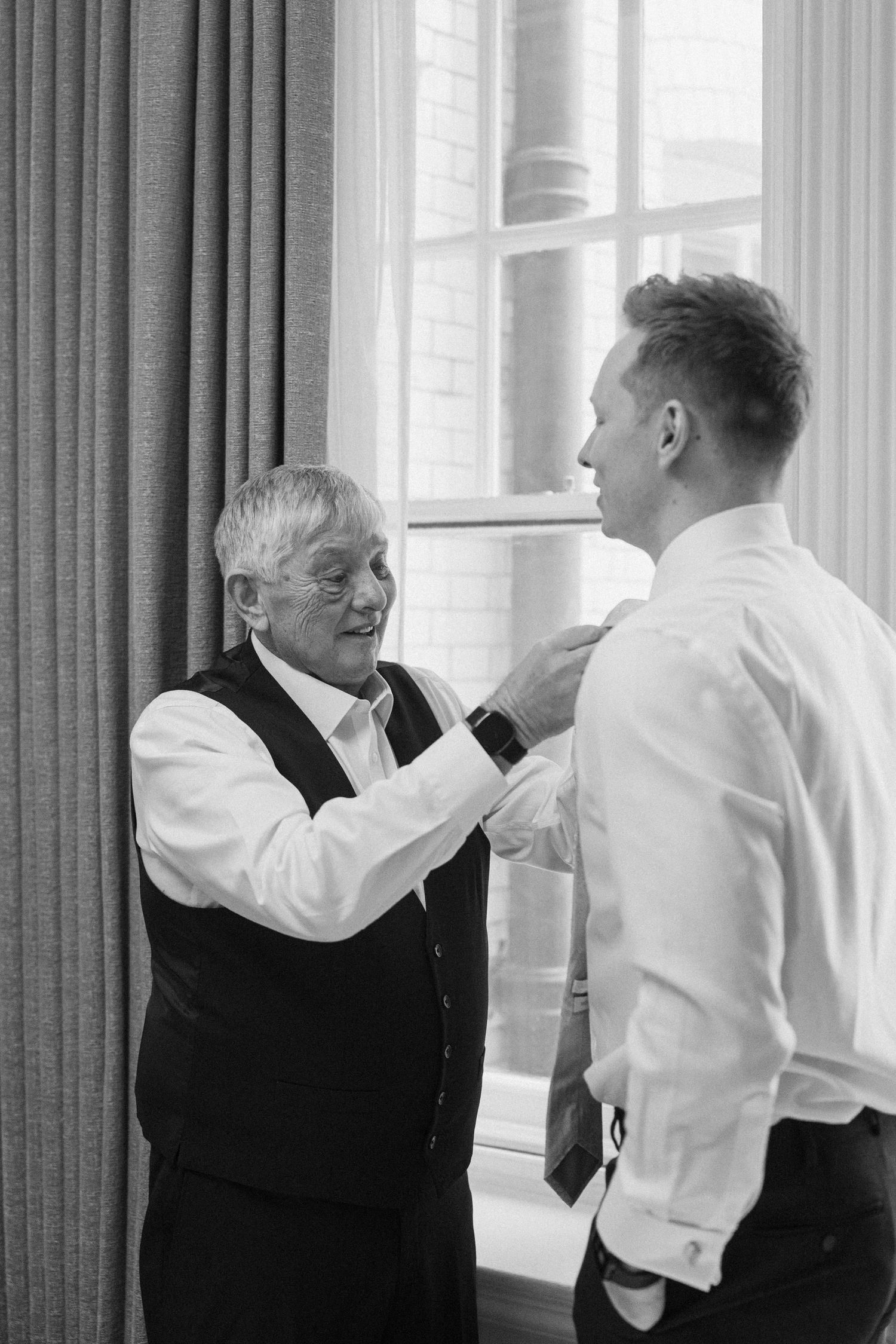 An older man smiles while adjusting a young grooms tie in a bright room in a black and white wedding photo.