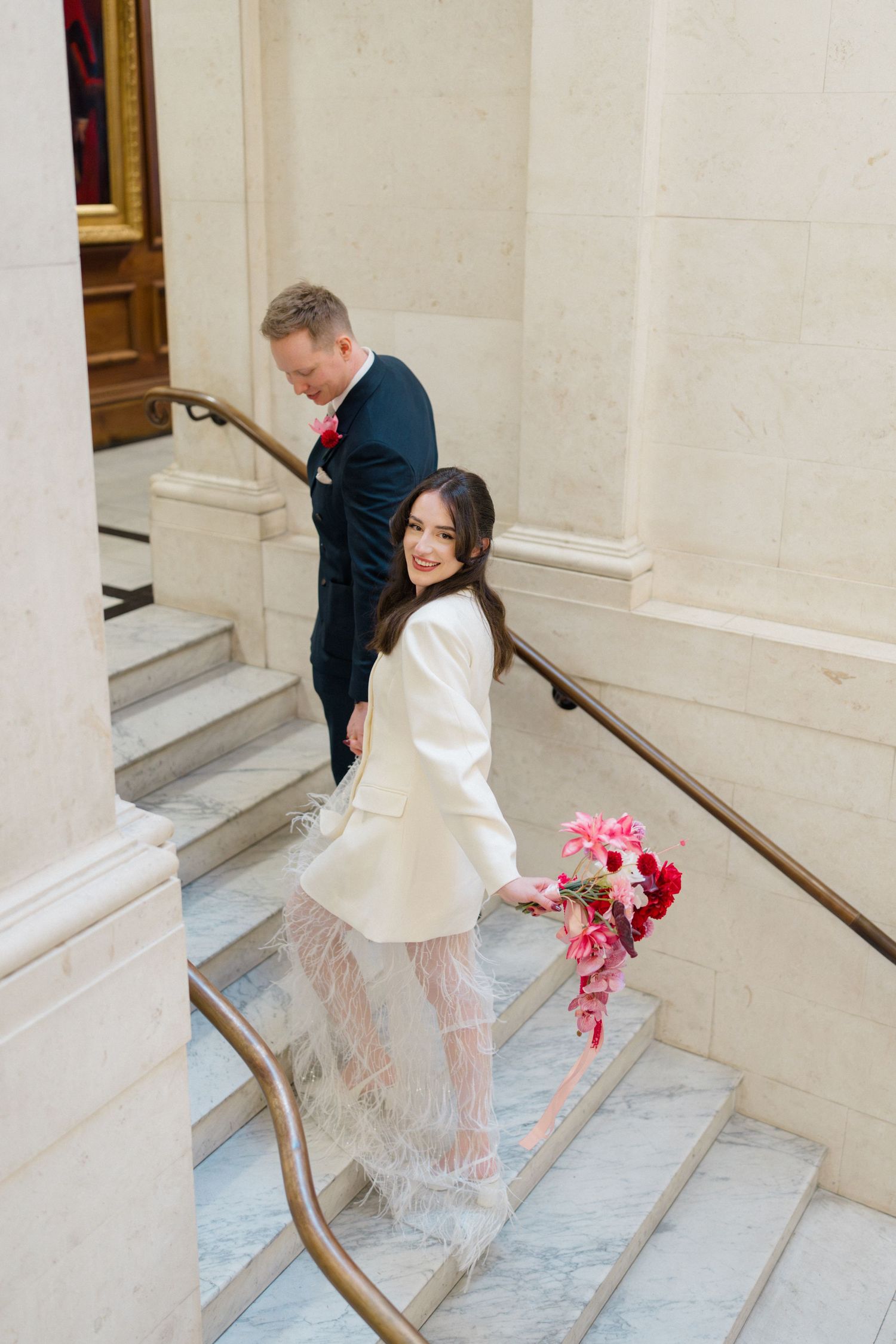Bride in white feather wedding outfit smiles on marble staircase holding pink bouquet.