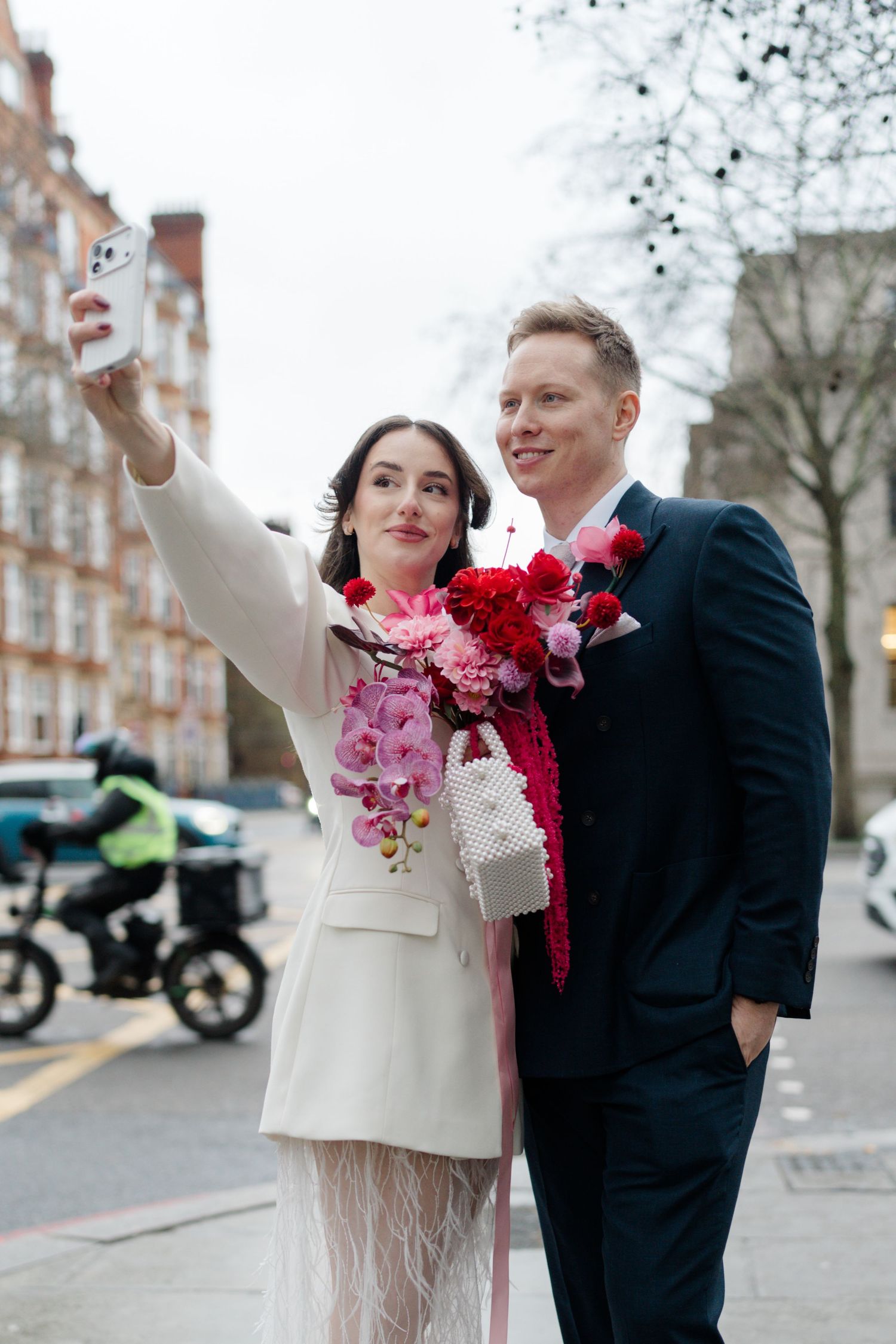 Bride and groom taking selfie on city street, bride holding vibrant pink and red floral bouquet in white feather skirt.