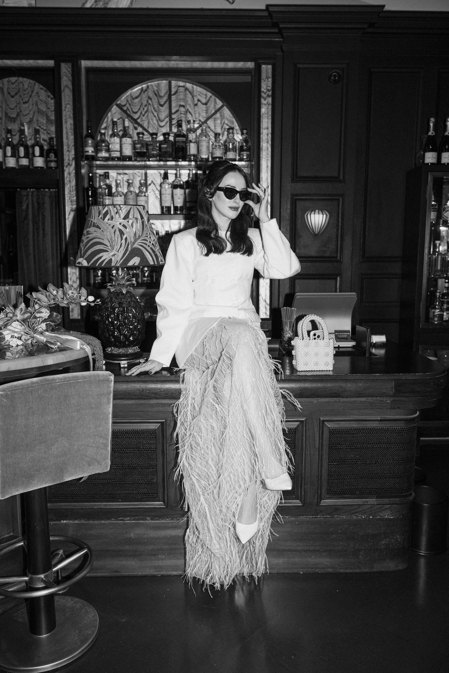 Black and white photo of bride in white feather trousers posing at luxury hotel bar.