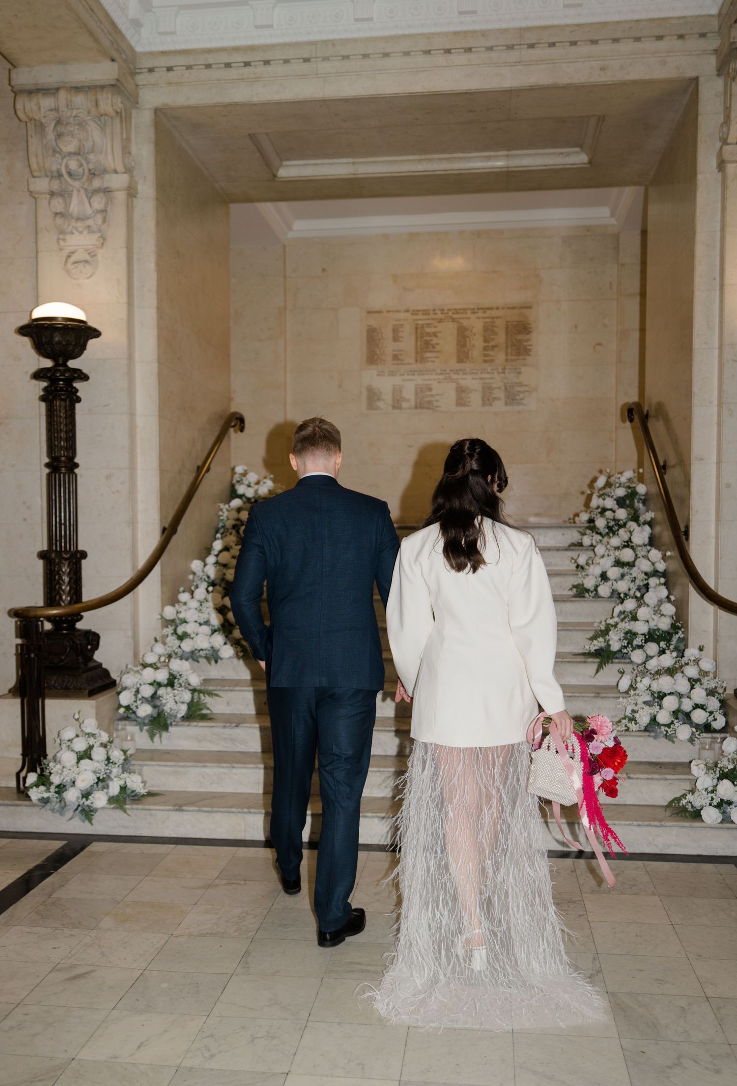 Bride and groom walk hand in hand toward floral-decorated staircase in elegant wedding venue.