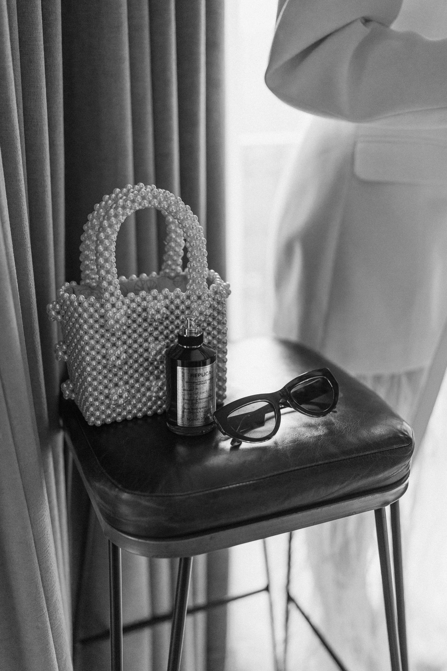 Black and white flat lay of a beaded handbag, sunglasses, and perfume bottle on a leather stool.