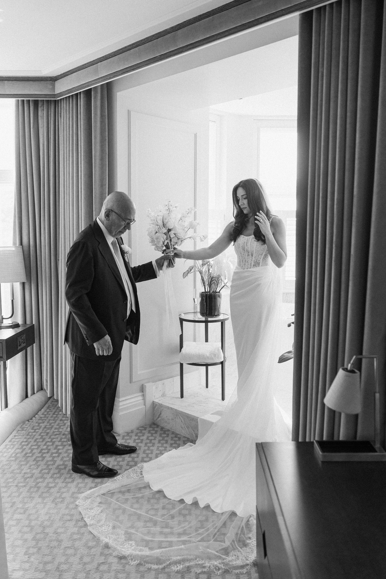Black and white photo of a father handing his daughter her bridal bouquet as she prepares in her wedding gown.