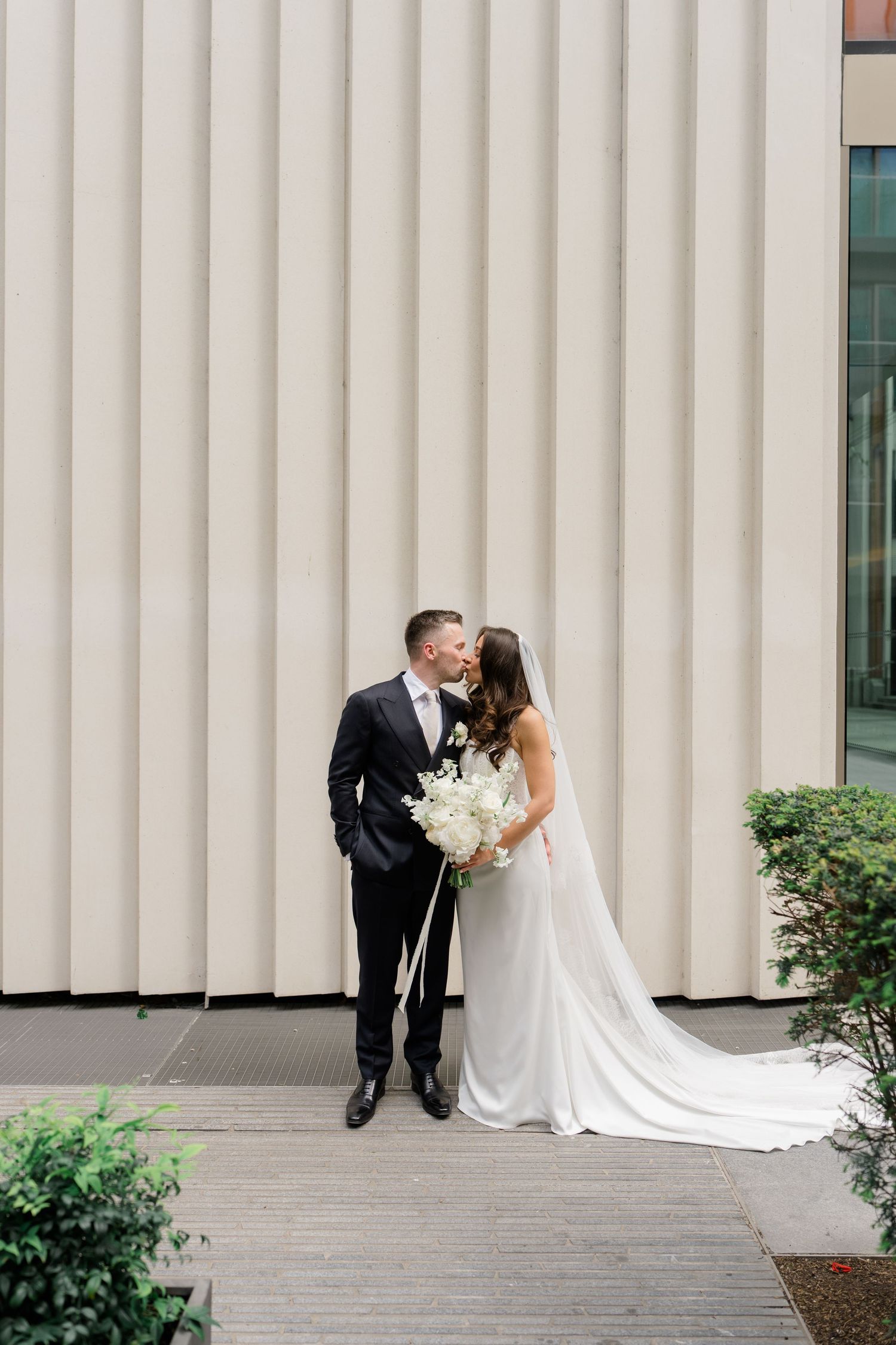 Bride and groom share a kiss outside modern building, bride holding white floral bouquet with flowing train.