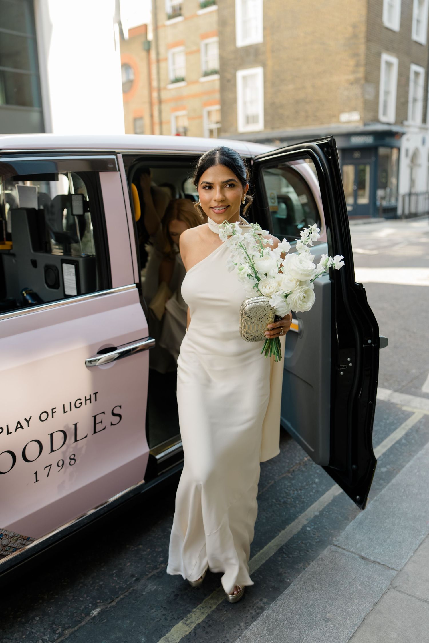 Radiant bride in a white satin gown stepping out of a pink Boodles branded London taxi holding a white floral bouquet.