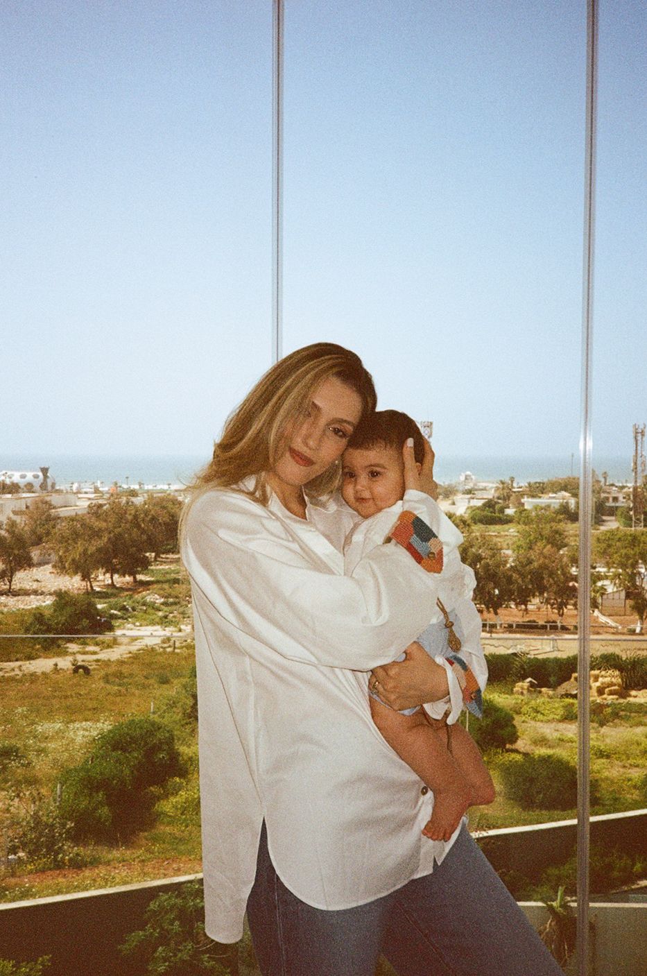 Woman in white shirt holding a baby near glass windows with green landscape and ocean view in background.