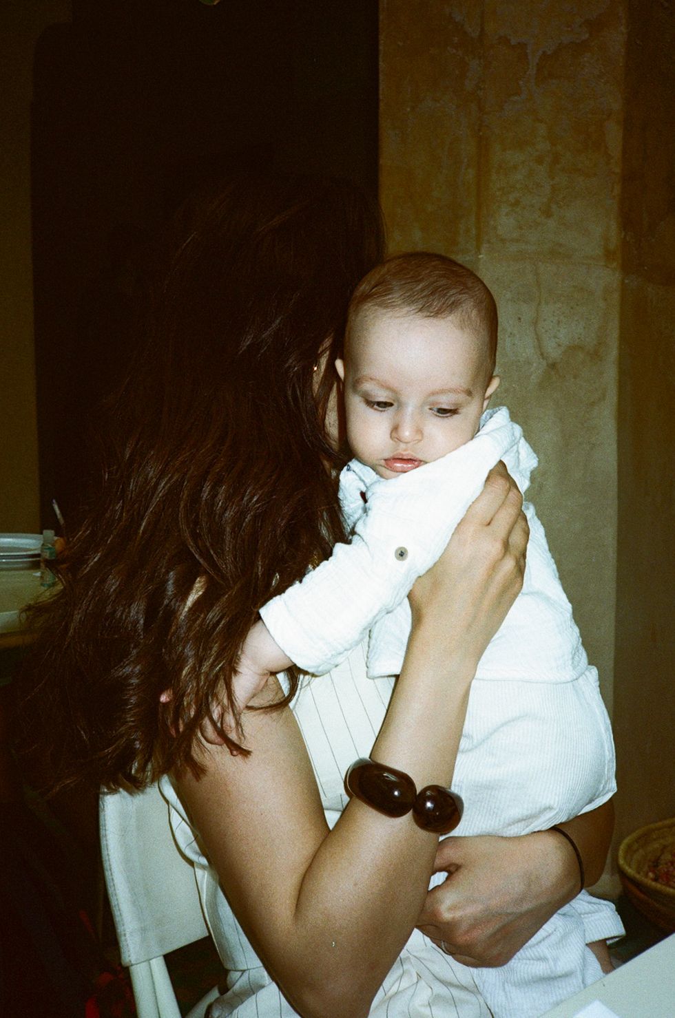 A mother with dark hair holds her baby dressed in white, gently cradling the infant close to her chest.