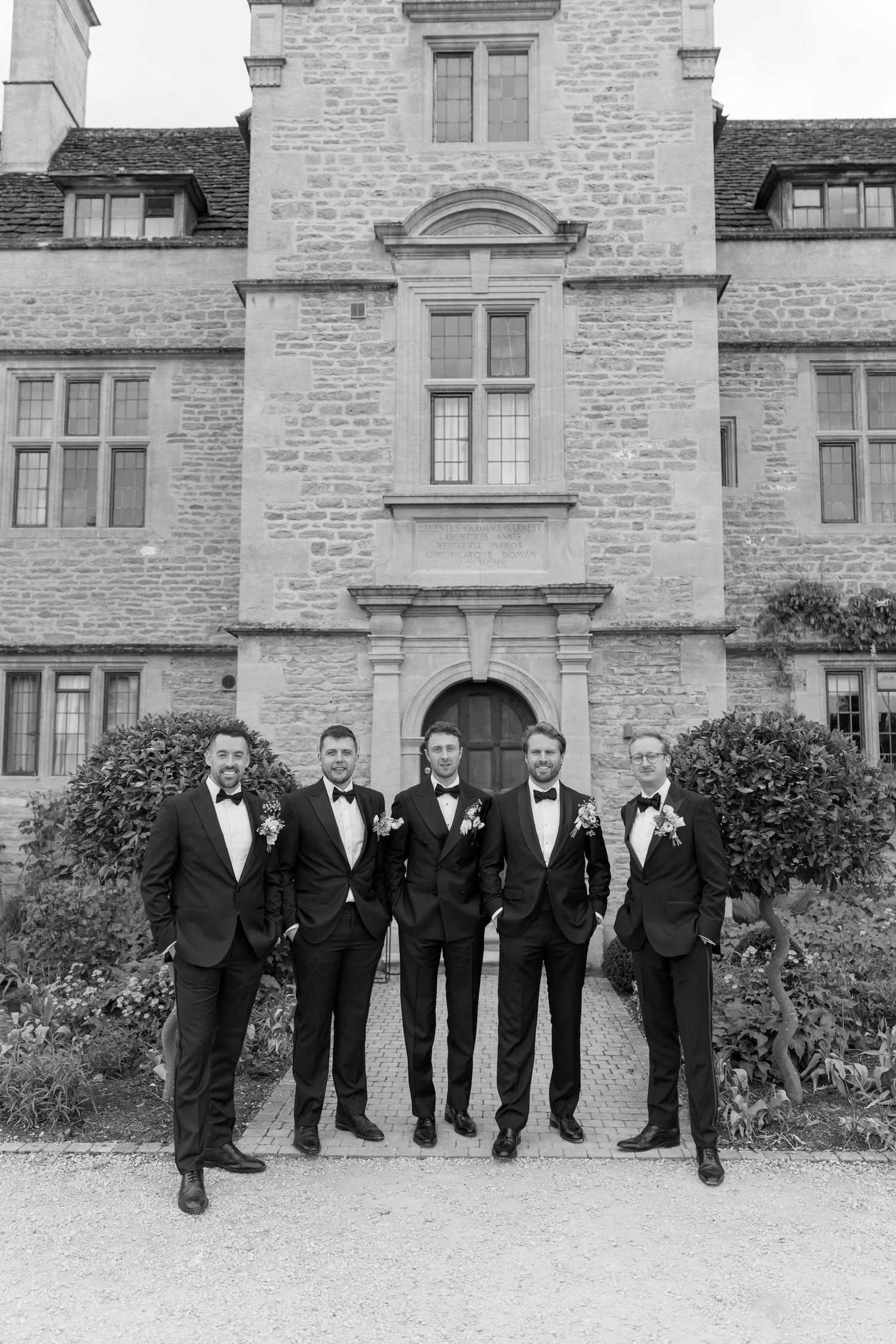 Five groomsmen in black tuxedos posing before historic stone manor house in black and white.