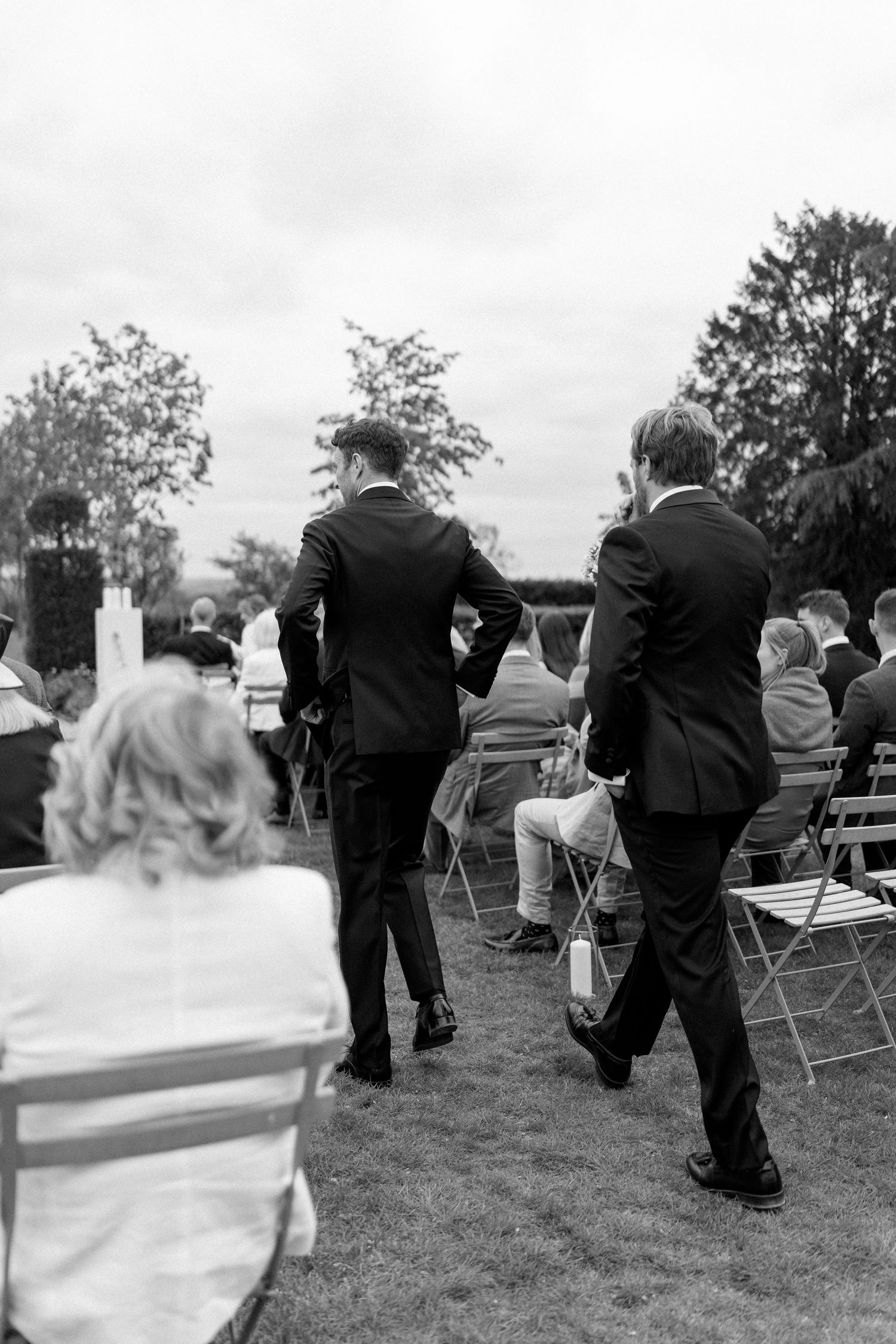 Two groomsmen in black suits walk down outdoor wedding aisle in black and white photo.