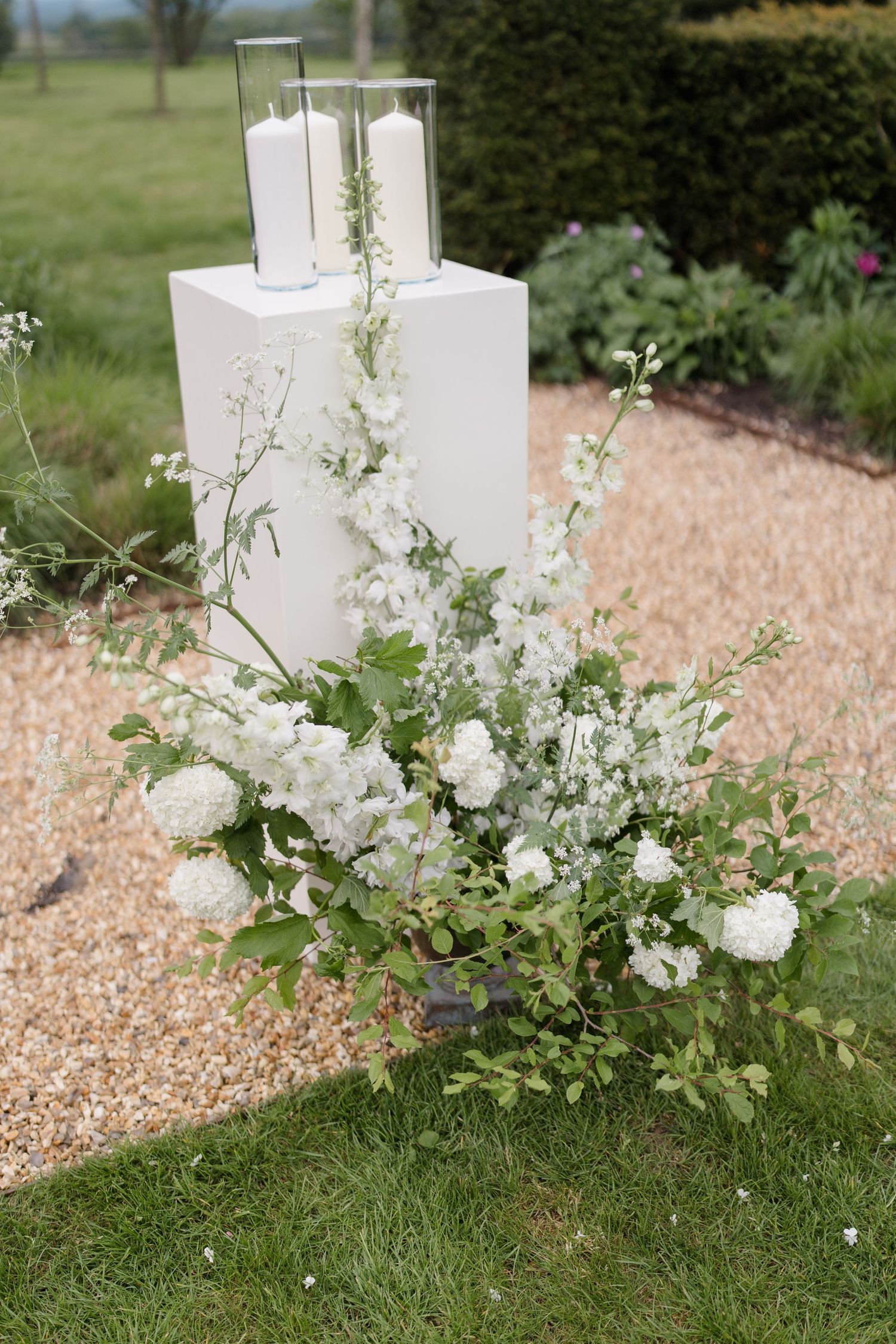 White floral arrangement with roses and greenery beside white pedestals with pillar candles at outdoor wedding.