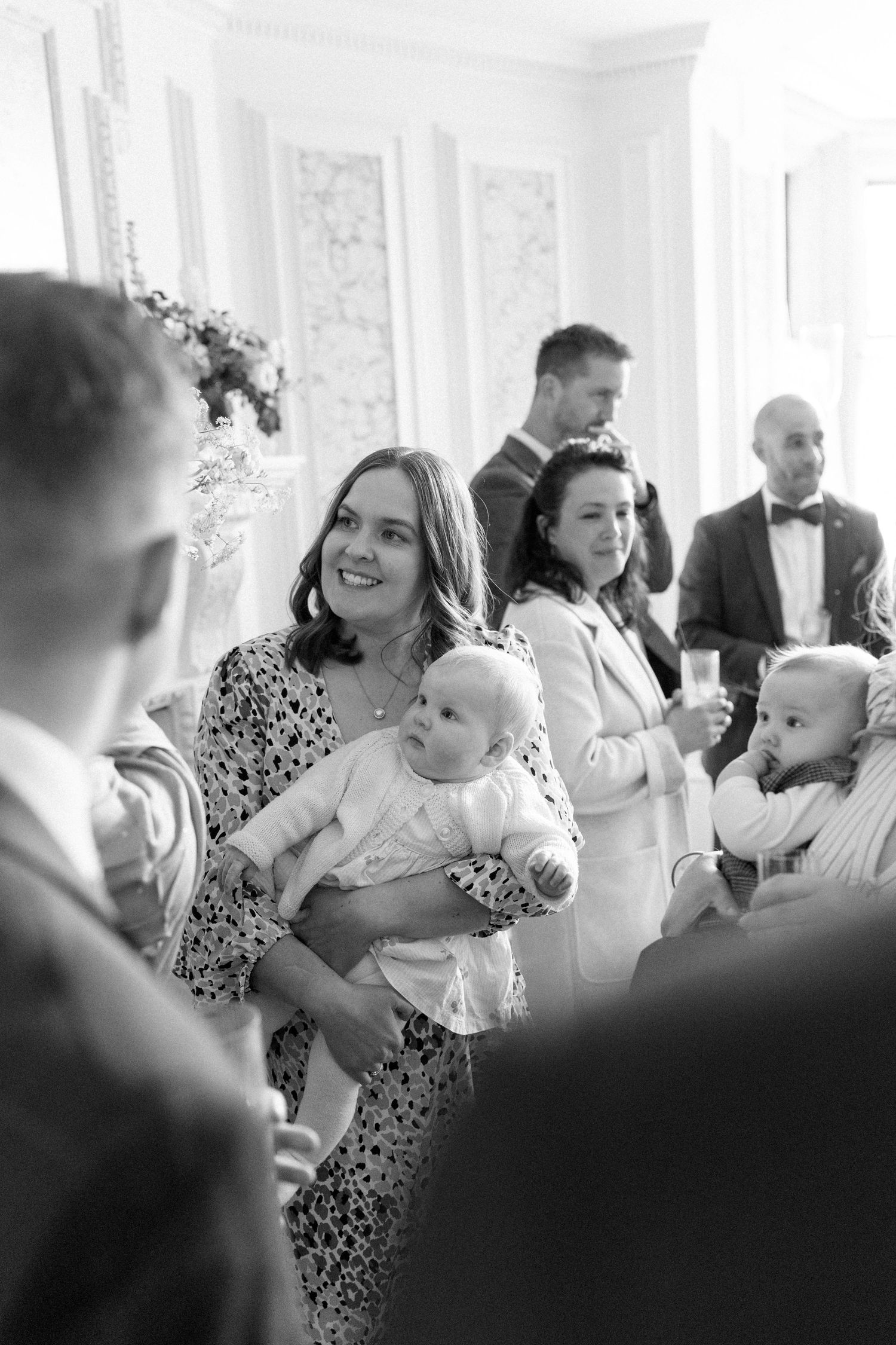 Wedding guest smiles while holding baby at elegant indoor reception in classic black and white photo.