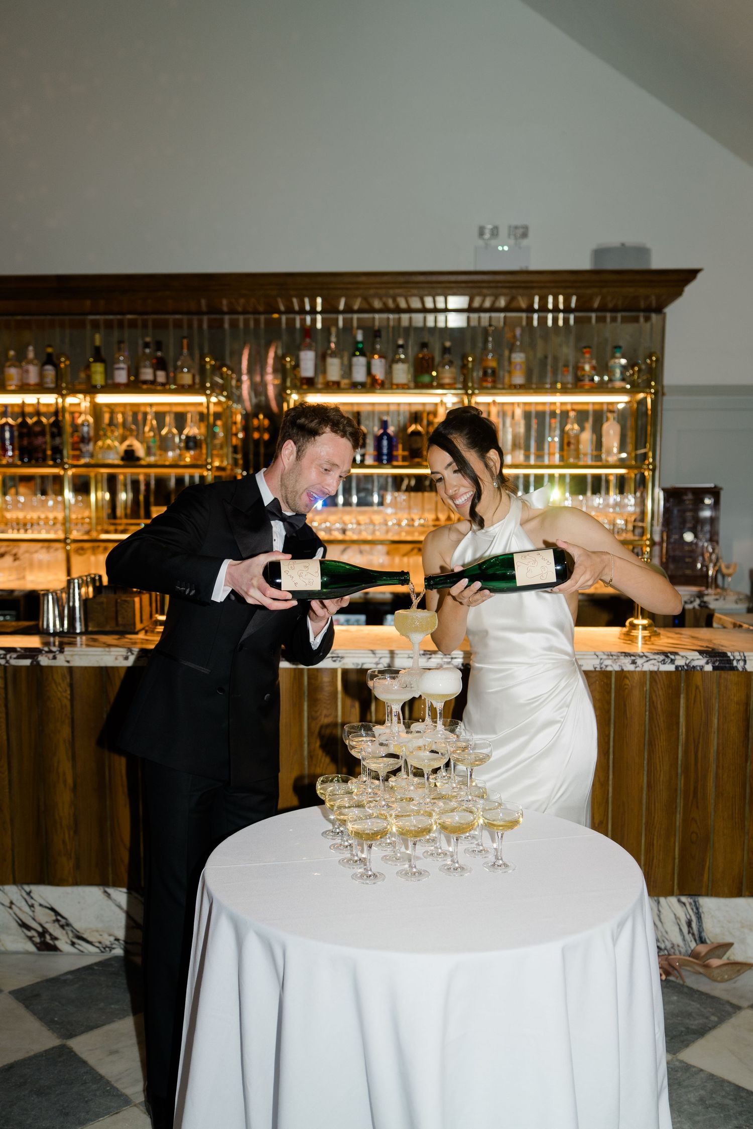 Bride and groom pour champagne together into an elegant tower of coupe glasses at reception.