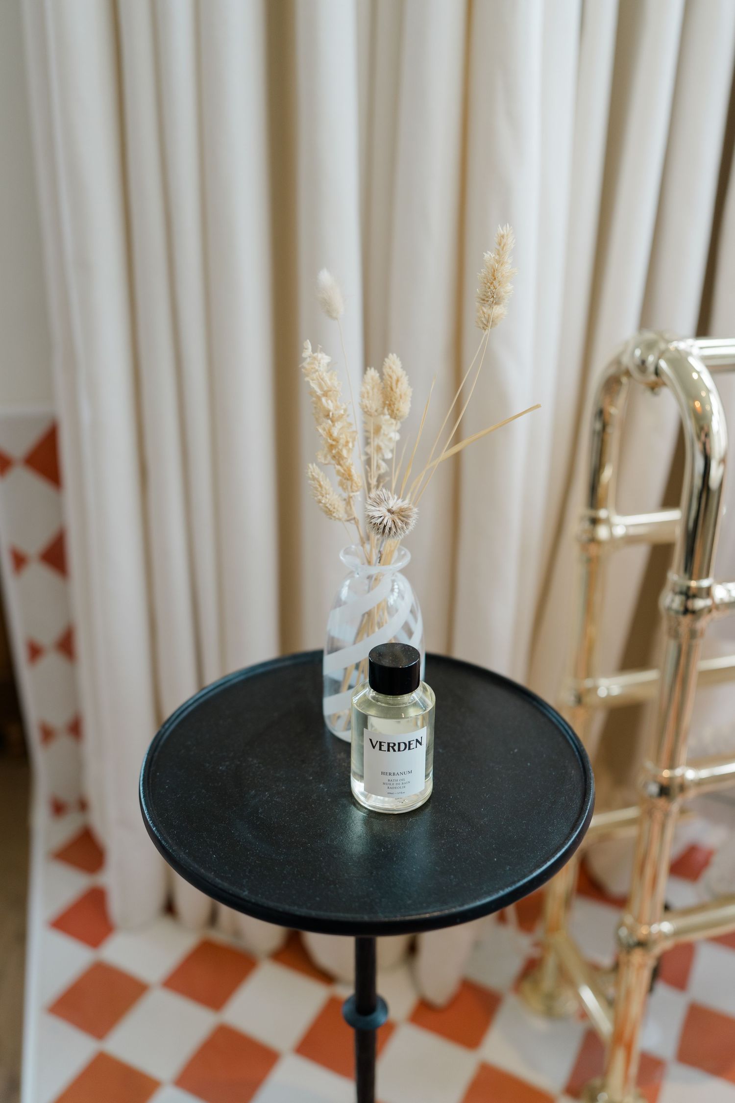 Verden perfume bottle with dried flowers on a black side table beside a gold towel rail.