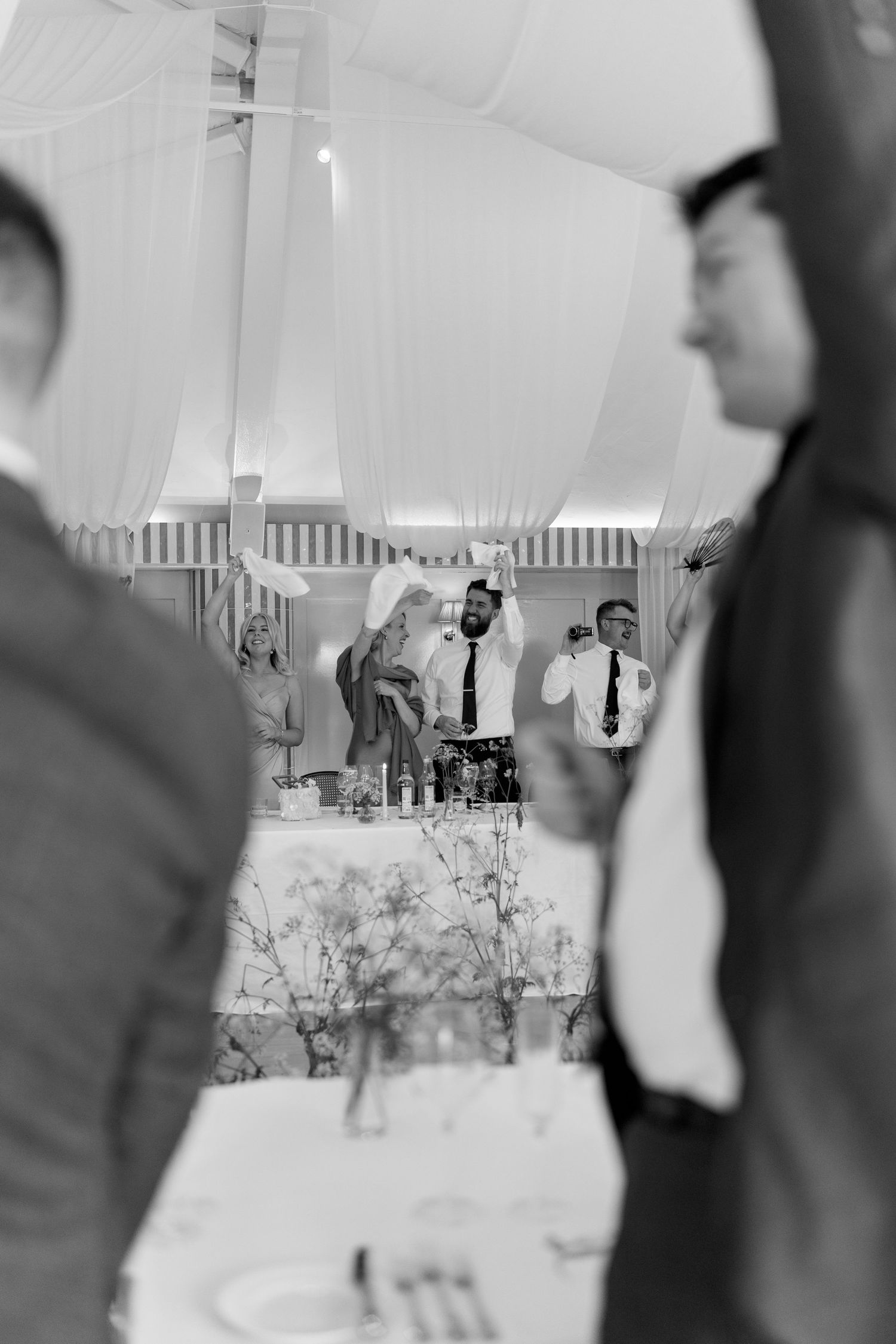 Wedding guests celebrating during reception speeches in elegant draped marquee in black and white.