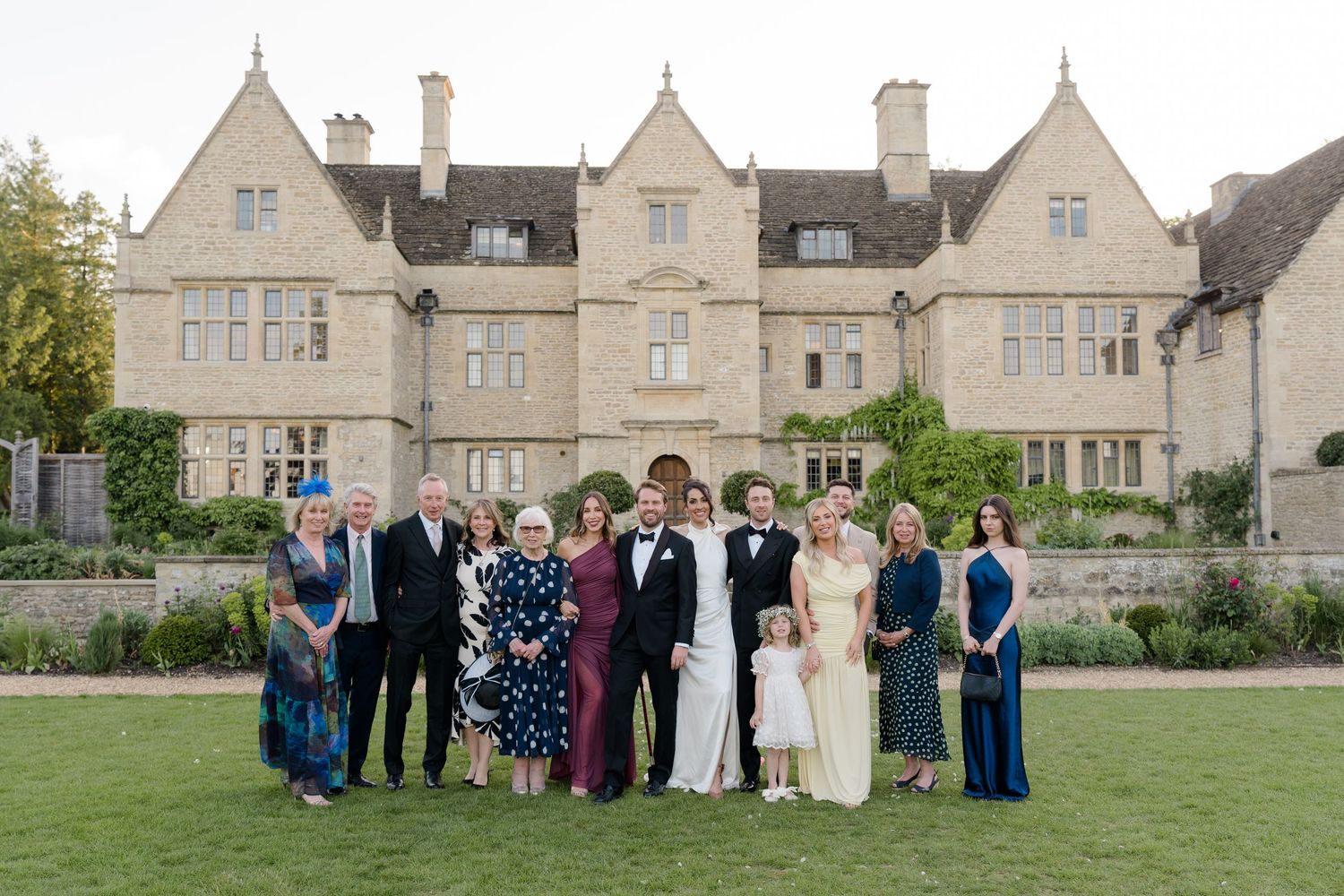 Wedding couple and extended family pose for formal portrait on lawn in front of grand stone country manor.
