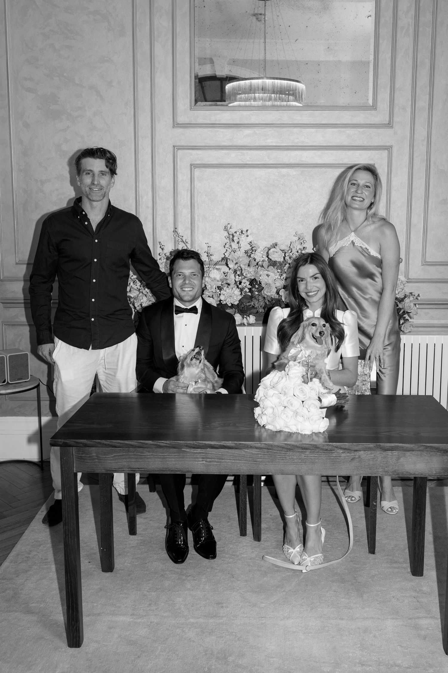 Wedding party poses at signing table with dogs and floral arrangements in elegant room.