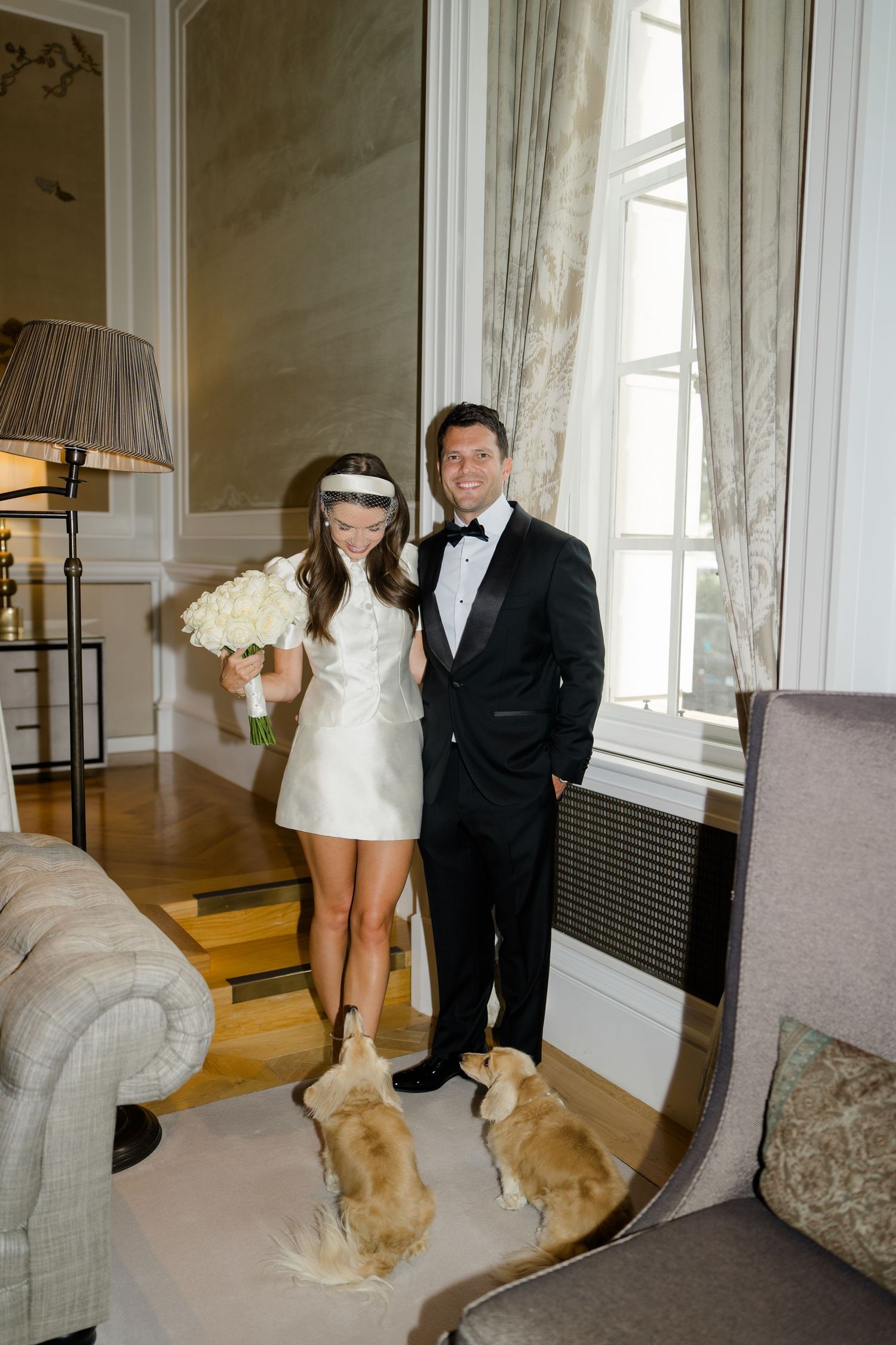 Bride in white mini dress holds bouquet while groom in tuxedo poses with two dachshund dogs in elegant hotel suite.