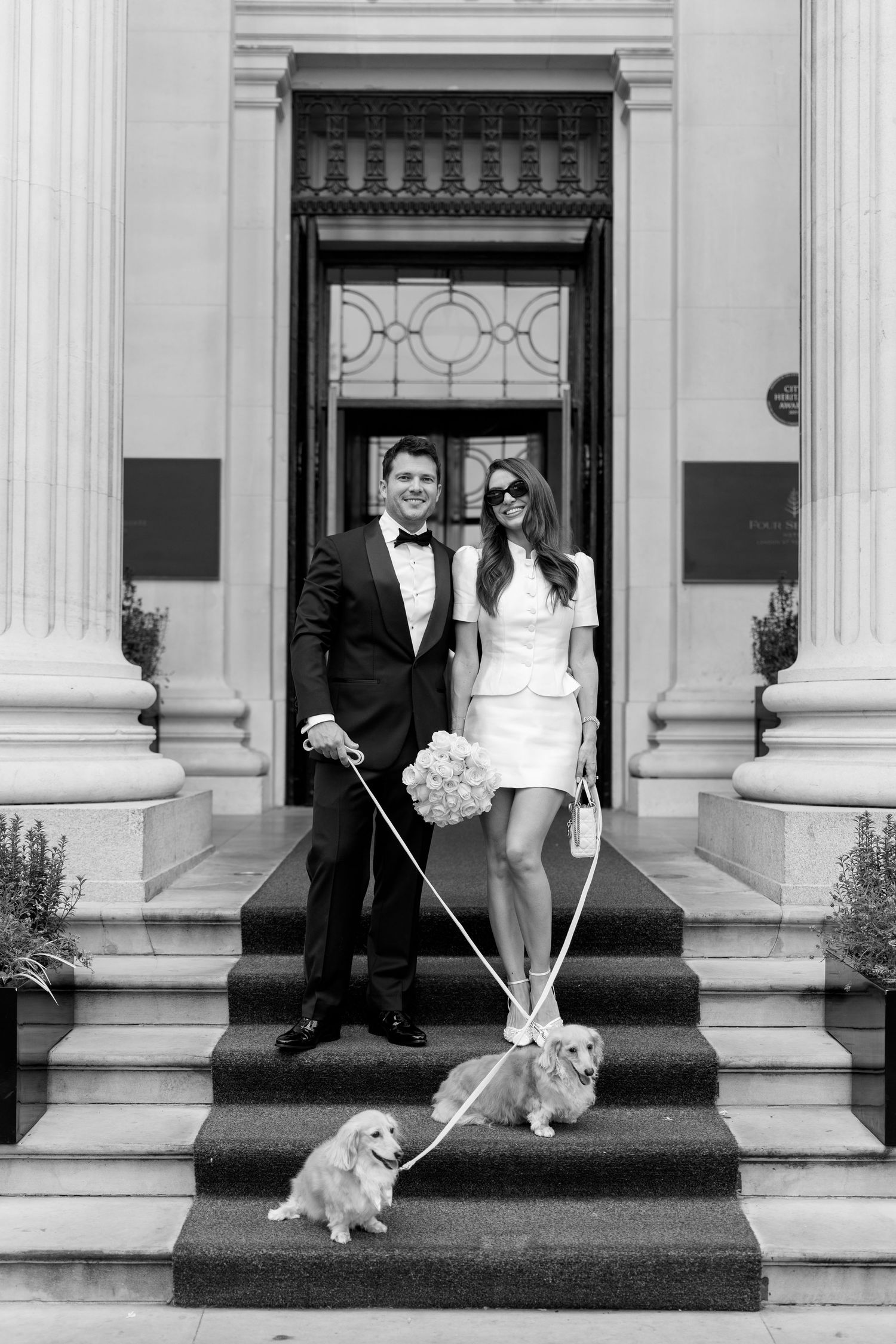 Elegant couple in wedding attire pose on steps with two golden dogs on white leashes.
