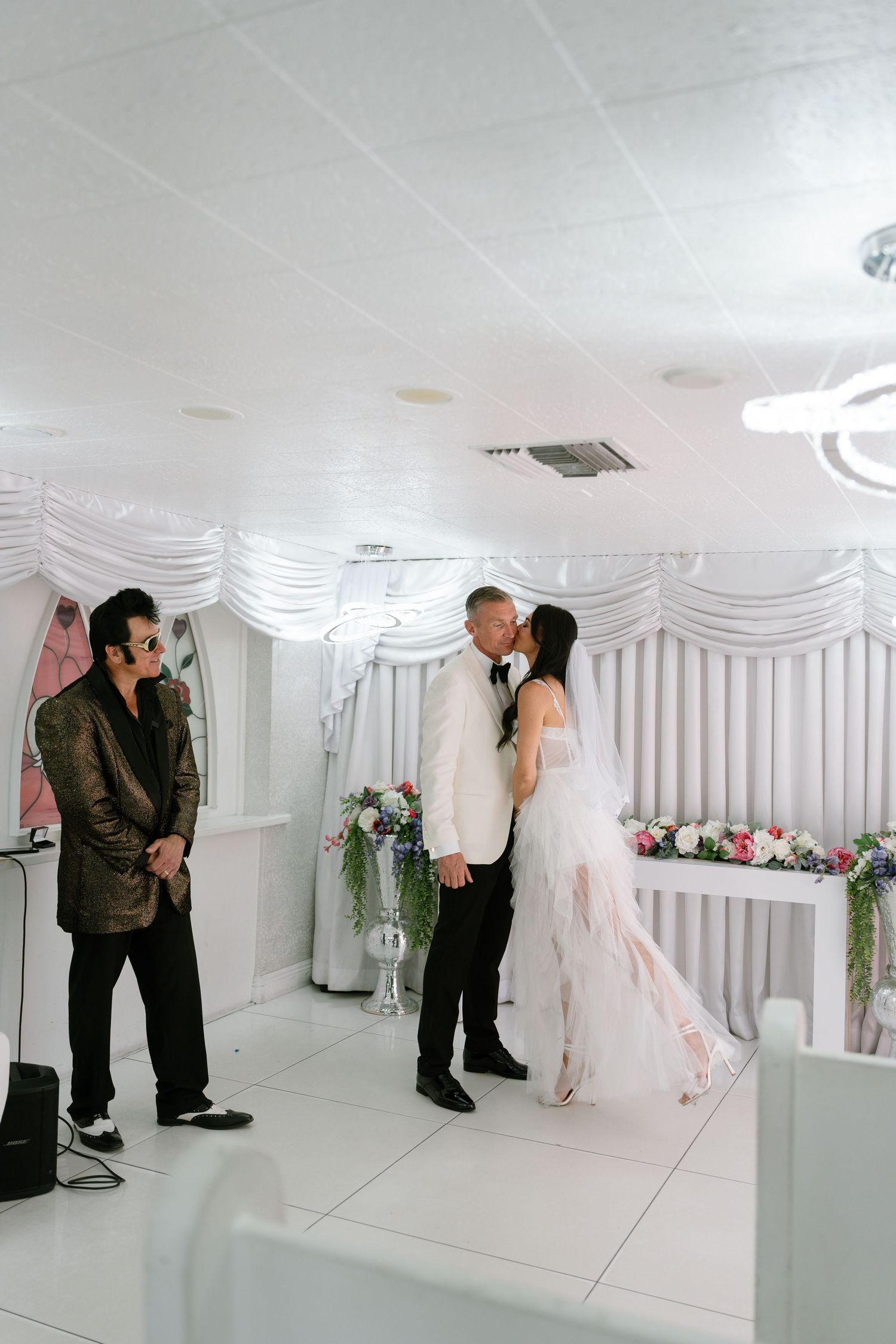 Wedding ceremony in progress with an officiant and couple in a white-draped chapel setting.