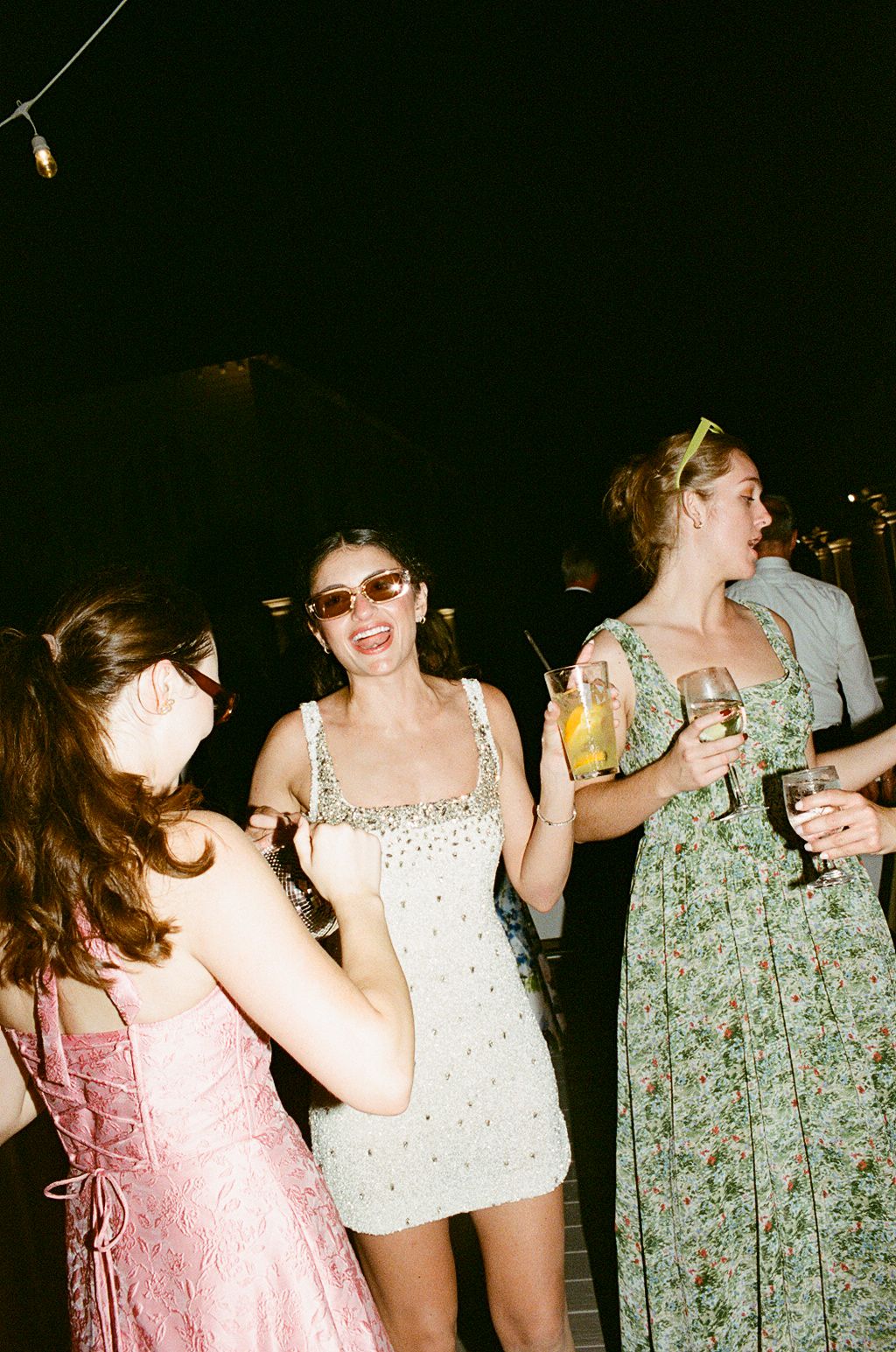 Three young women in elegant dresses dancing and holding drinks at a nighttime party event.