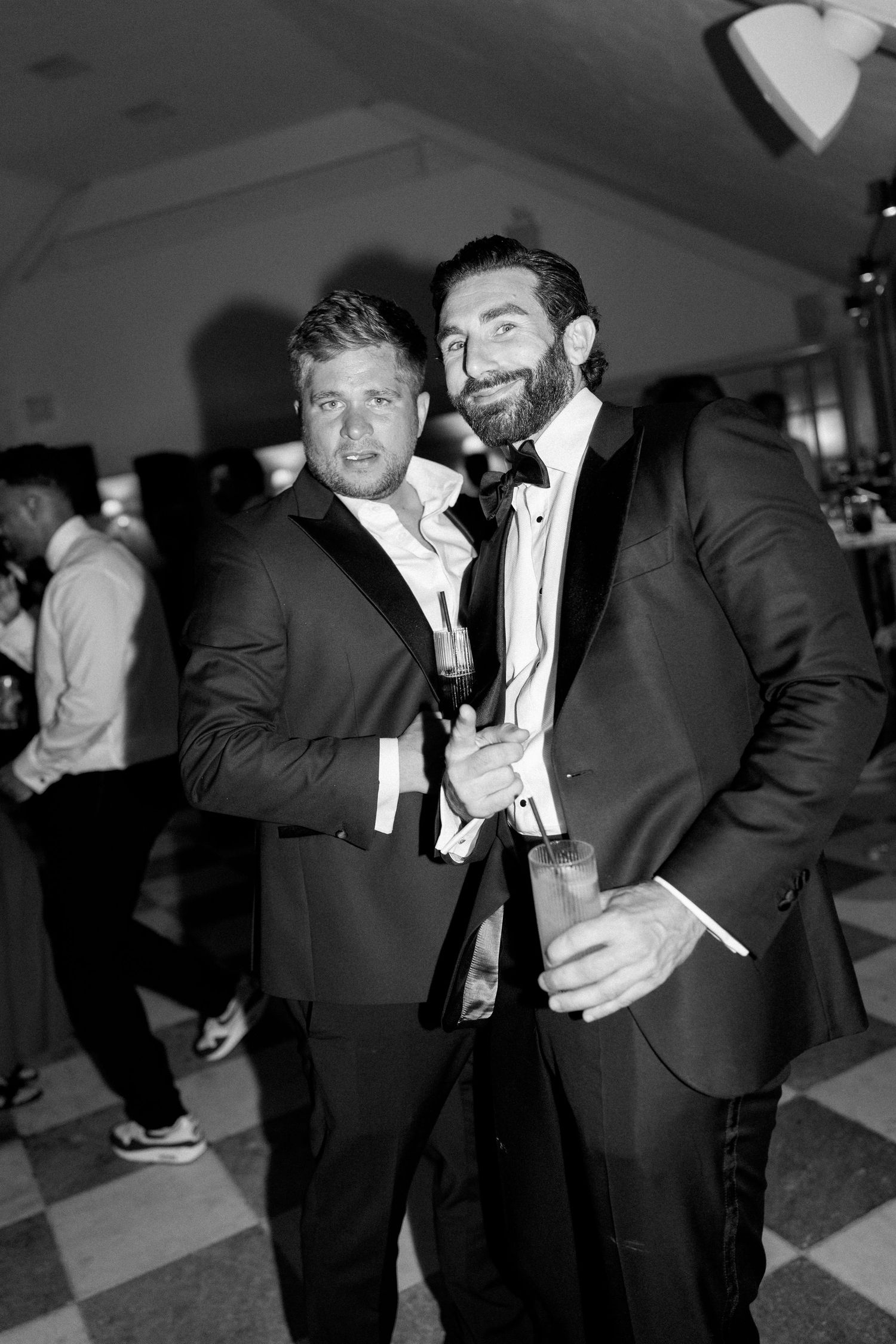 Black and white photo of two men in tuxedos smiling and posing together at a wedding reception.