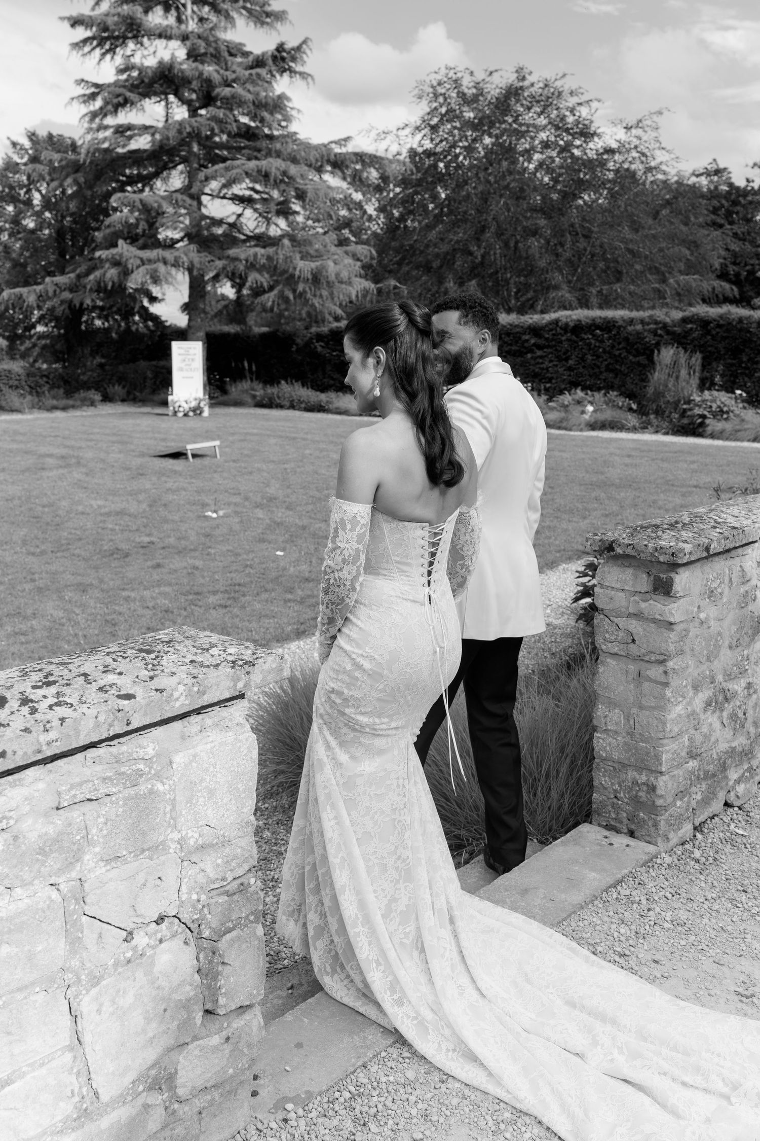 Black and white photo of bride in lace corset gown and groom in white jacket at stone garden wall.
