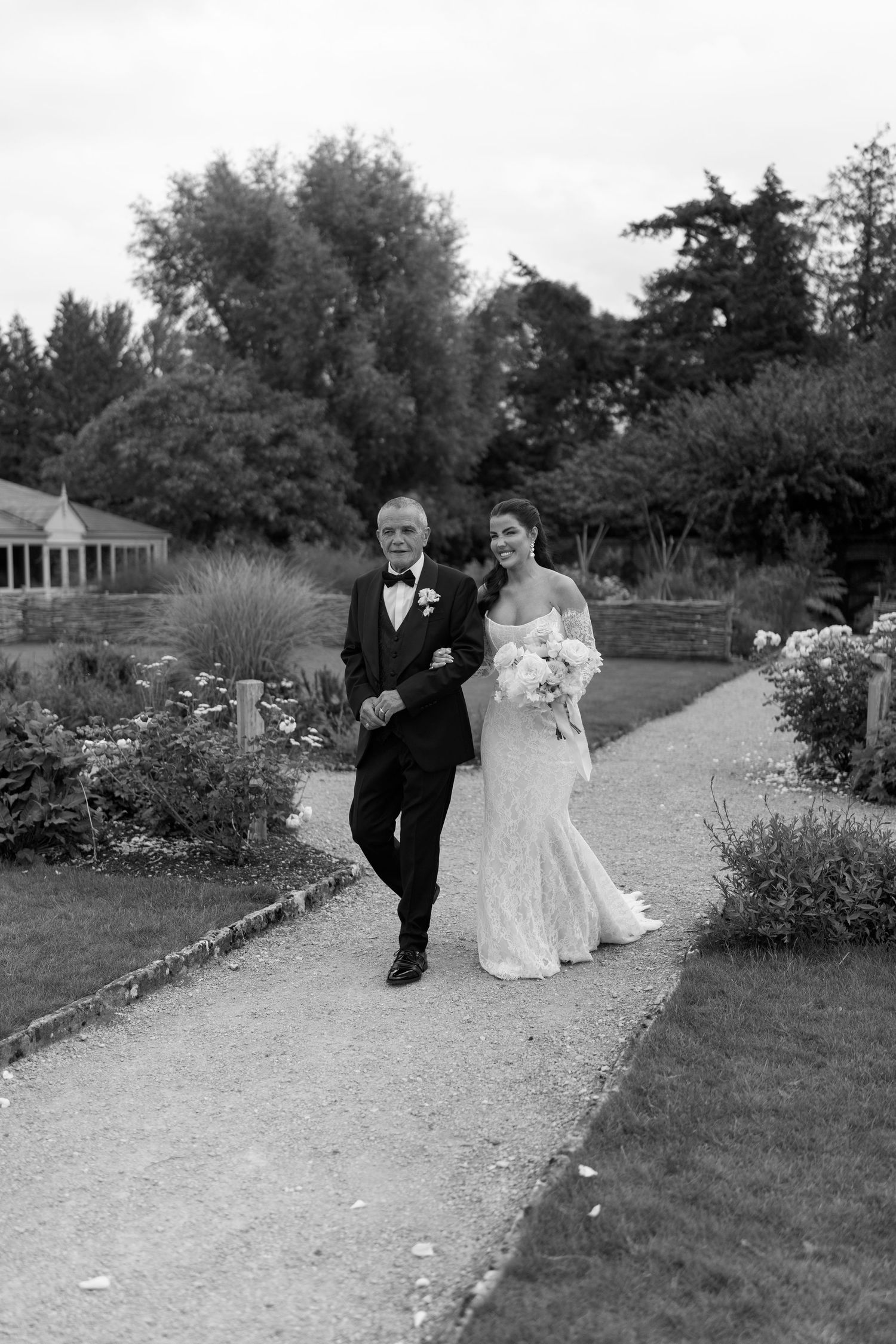 Bride in fitted lace gown walking with her father through a garden path in black and white.